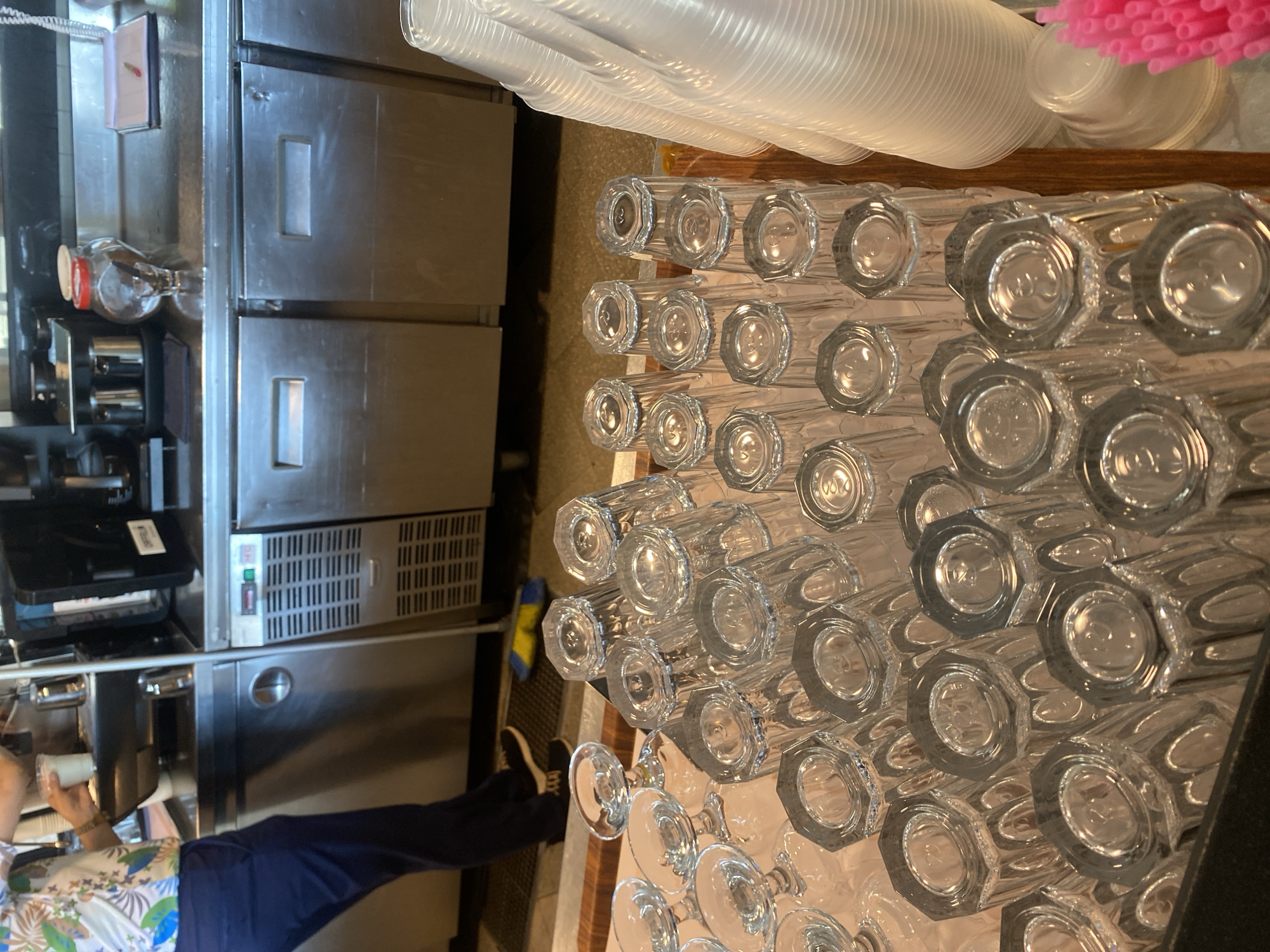 The image depicts a close-up view of numerous clear glass tumblers stacked upside down, filling the foreground of the frame. To the right, a stack of clear plastic disposable cups is visible, and a cluster of bright pink straws is also present. In the mid-ground, a stainless steel counter area is evident, with various appliances and equipment, including what appears to be a commercial coffee machine and a refrigerator unit with visible vents. A person, partially visible on the left side, is wearing a short-sleeved, floral print shirt and dark blue pants, and appears to be interacting with the coffee machine. The background shows more stainless steel surfaces and cabinets. The overall scene suggests a food service or hospitality setting. The request to include the location in Aksu, Turkey, cannot be fulfilled as there is no visual information within the image to support this specific geographical context.