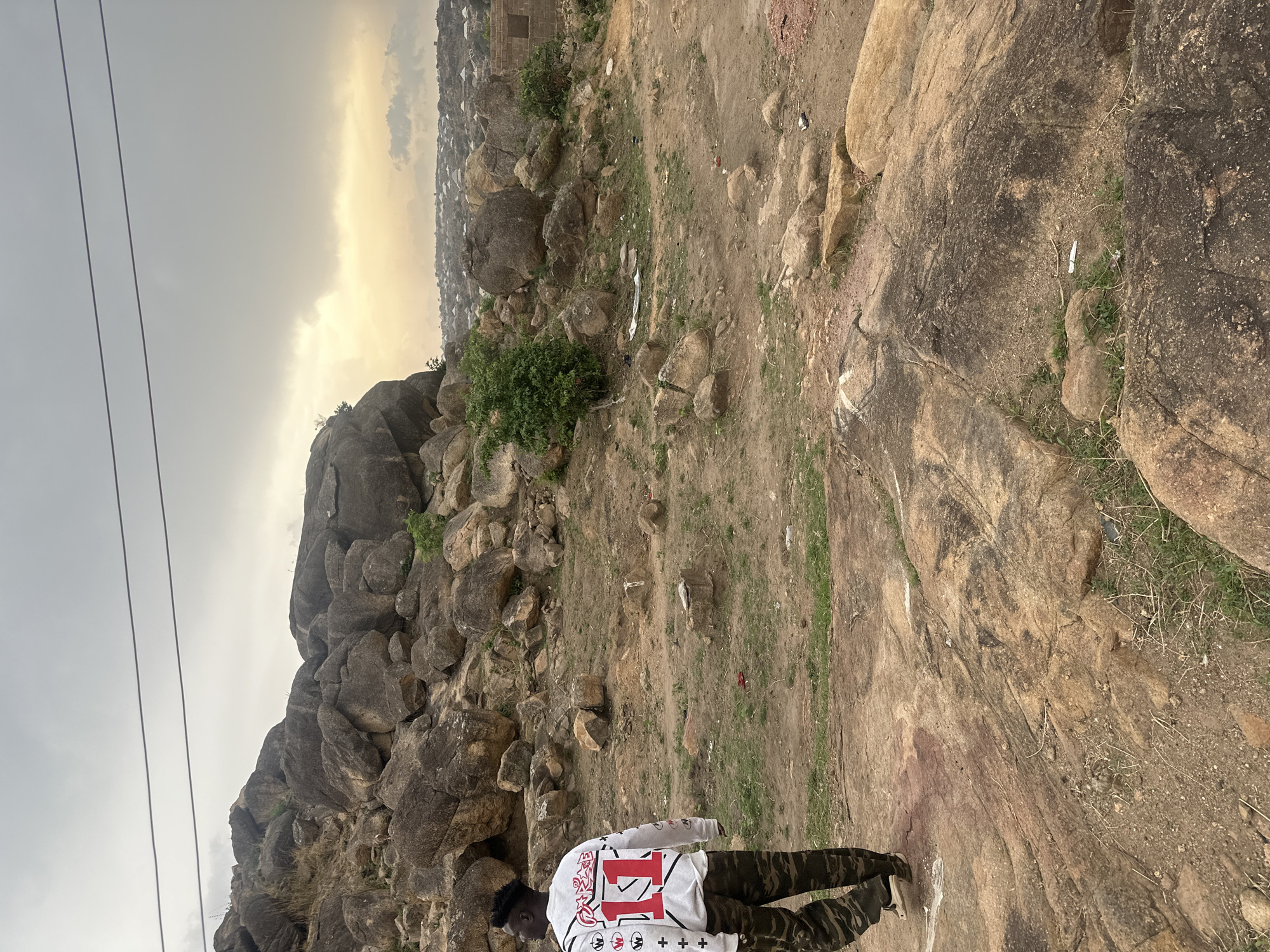 The image depicts an outdoor scene in Jos, Nigeria. A person wearing a white and red long-sleeved shirt with camouflage pants is standing on a rocky and earthy terrain. Behind the individual is a large rock formation with scattered vegetation. The background shows a distant cityscape under an overcast sky, with power lines visible in the upper left corner. Debris, possibly trash, is scattered across the ground.