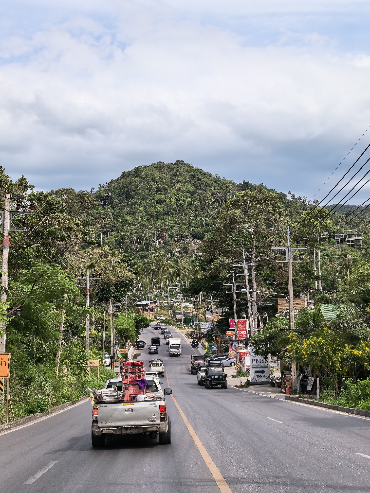 This is a vertical, eye-level, full shot of a road in Ko Samui District, Thailand. The road curves slightly upward and to the left, leading towards a densely forested hill. On the right side of the road, there are several businesses, including one with a sign reading "PBISS International School". Utility poles with numerous wires run along the side of the road. The sky is partly cloudy with large white clouds. The road is paved with asphalt and has a yellow dividing line. Several vehicles are visible on the road, including a white pickup truck in the foreground carrying what appears to be stacks of red boxes and a purple tarp. Other vehicles on the road include cars, a black SUV, and a white truck. The vegetation is lush and green, with palm trees and other tropical foliage. The overall impression is of a busy road in a tropical, hilly area.