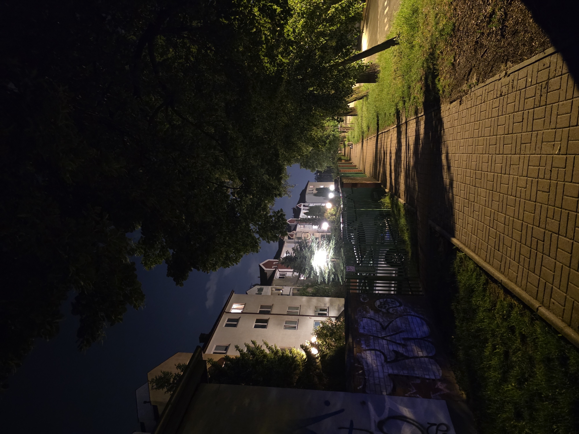 At night, a street in Warsaw, Poland, is depicted with trees overhead. The sidewalk, made of brown bricks, runs along a green fence. A building with lit windows is on the left. The sky is visible between the trees and buildings. Graffiti is seen on a wall along the sidewalk. Street lights illuminate the scene.