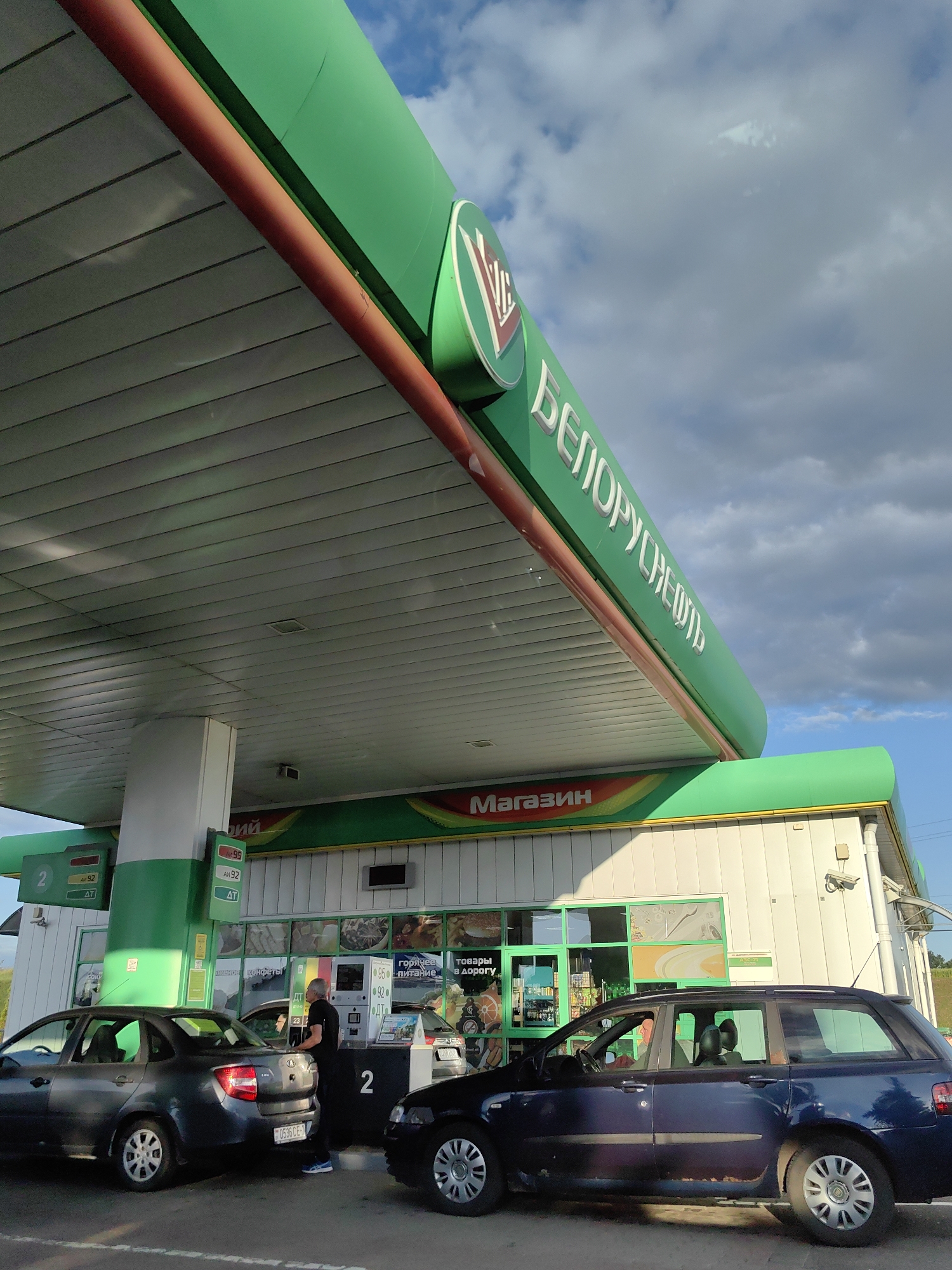 At a gas station in Фаніпальскі сельскі Савет, Belarus, a dark gray sedan is parked to the left of a gas pump with its rear facing the camera. A man in a black shirt and blue shoes is standing at the pump. In front of him, a dark blue station wagon is parked, with its driver visible inside. To the right of the station wagon, the front of a black car is visible. The gas station has a green canopy with the white lettering "БЕЛОРУСНЕФТЬ" visible on it, and "Магазин" in orange letters above the store. The sky is partly cloudy.