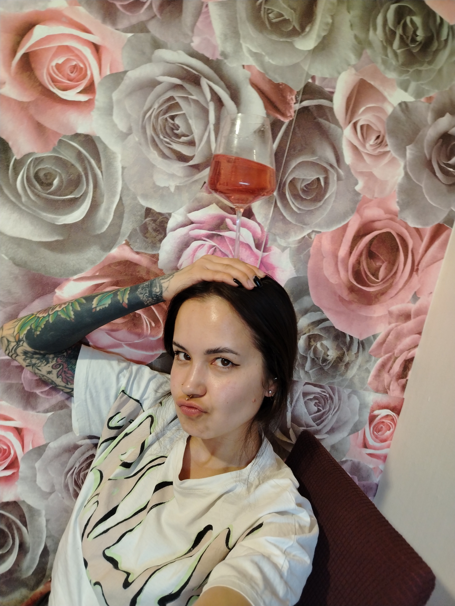 A medium shot depicts a woman indoors, holding a glass of red liquid on her head. She has dark hair, fair skin, and a nose ring. Her left arm, visible, features full sleeve tattoos. She is wearing a white t-shirt with a black and beige design. Behind her, a wall is covered in a pattern of pink and gray roses. A maroon-colored sofa cushion is visible to her right. The city of Noviy, Russia is not discernible from the image.