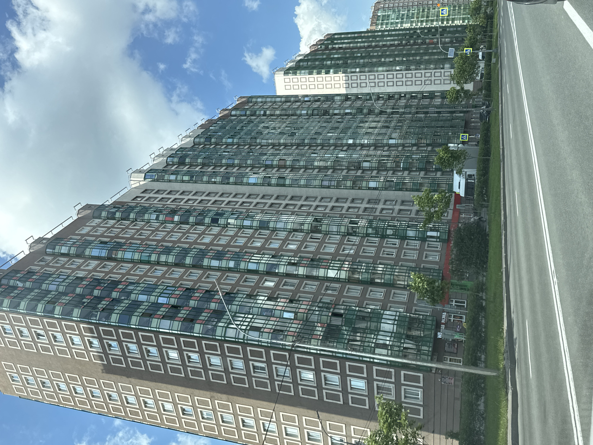 The image displays a long, modern multi-story building situated alongside a multi-lane asphalt road, under a bright blue sky dotted with scattered white clouds. The building, which occupies the left side of the frame, features a distinctive architectural style with alternating sections of dark-colored facade and reflective glass panels. Many parts of the building have rows of square windows, some framed in white, while other sections are covered by extensive glazing, possibly for balconies or large windows. The building's upper levels appear to be stepped or terraced.
A verdant strip of grass, dotted with small trees and bushes, separates the building from the road. The road is clearly marked with white double solid lines. Several yellow diamond-shaped road signs, featuring black pedestrian symbols, are visible along the roadside, indicating pedestrian crossings. A few vehicles are partially obscured by the vegetation.
Visible text on a sign near the building's base includes the letters "WB" and partially visible Cyrillic text "НАБ РАЗНА". The bright illumination suggests it is daytime with good weather. No people are prominently featured. The scene captures an urban or suburban setting in Poroshkino, Russia.