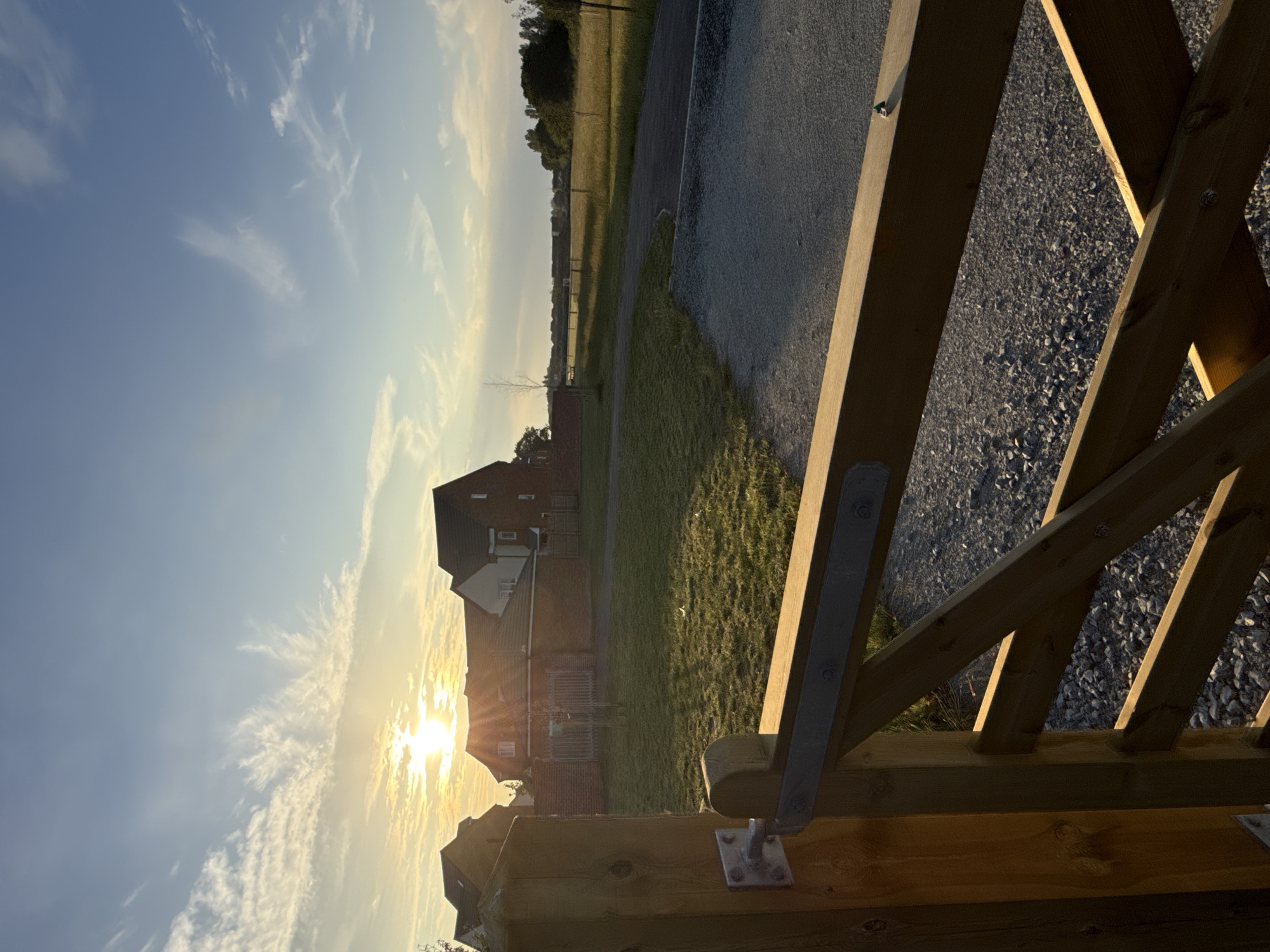 This image captures a serene sunset scene over a newly developed residential area in Great Yarmouth, United Kingdom. The sun, a bright orb low in the sky, casts a warm glow, illuminating wispy clouds and the facades of brick-built houses. The foreground is dominated by a rustic wooden gate, its natural wood grain and metal hinges clearly visible, leading onto a gravel path. Beyond the gate, a neatly manicured lawn stretches towards a paved pathway and a distant field bordered by trees. The overall impression is one of peaceful, early evening light settling over a quiet, suburban landscape.