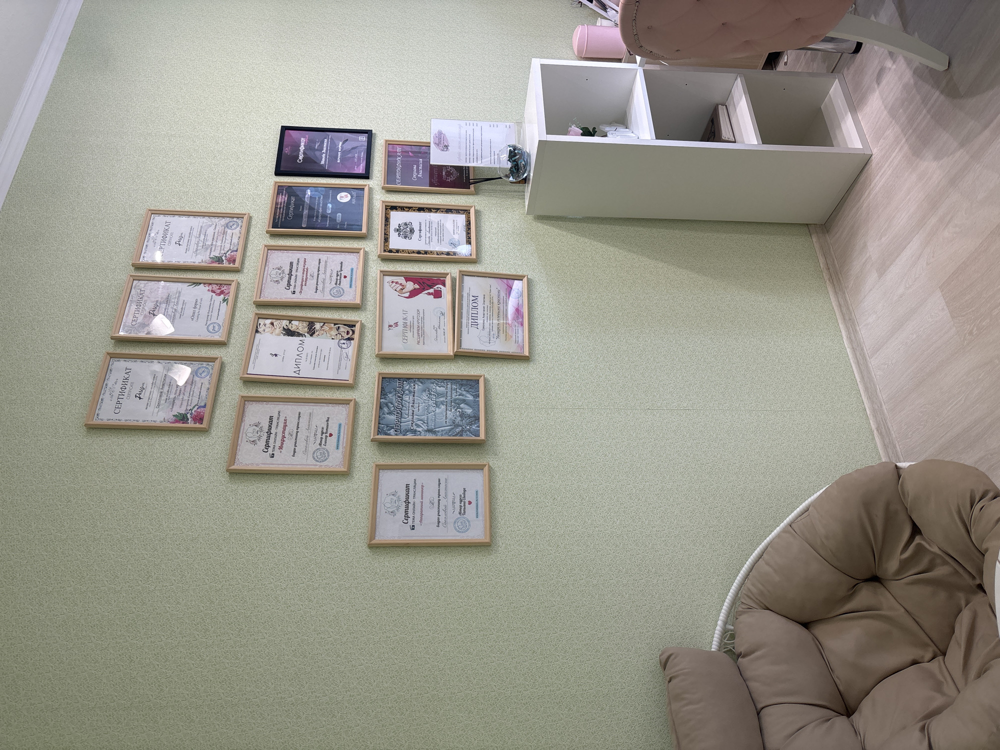The image shows a wall in what appears to be a beauty salon or office, adorned with numerous framed certificates and diplomas. The wall is light green with a subtle patterned wallpaper. The certificates are arranged in a somewhat haphazard, clustered fashion. Most of them are in wooden frames and appear to be related to beauty treatments or courses, indicated by the titles like "CERTIFICATE", "DIPLOMA", and words like "manicure". Some of the certificates display the name "Анастасия/Anastasia", suggesting they are for the same person.
Next to the certificates, a white shelving unit is visible, presumably for storing beauty-related products or supplies. Above this is a pink upholstered stool.
To the right of the wall is a beige cushioned chair. The flooring appears to be light-colored wood laminate.
The OCR text mentions "Ferma, Russia", "Соколова Анастасия Олеговна", which could be the person who acquired these certificates, and "Соколовой Анастасии", who is likely the same person mentioned on the certificates.