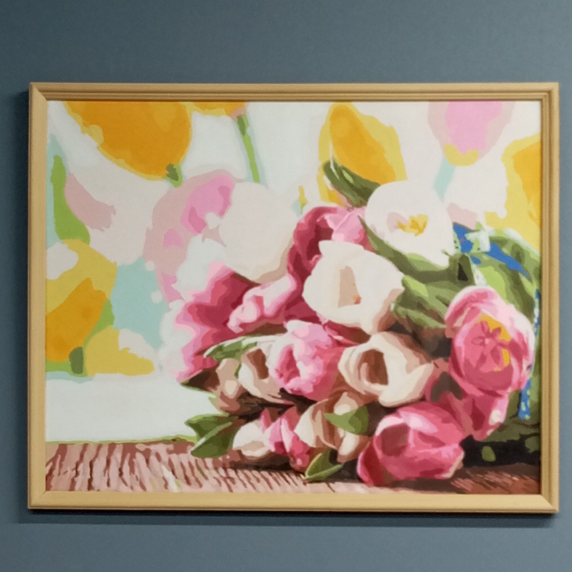 The image shows a framed painting of a bouquet of tulips. The tulips are primarily pink and white, with green leaves. The background consists of abstract shapes in yellow, pink, and light blue. The painting sits against a solid gray-blue wall. The location of the painting is unknown. It is not possible to conclude from this image it is in Pervomaysky, Russia.