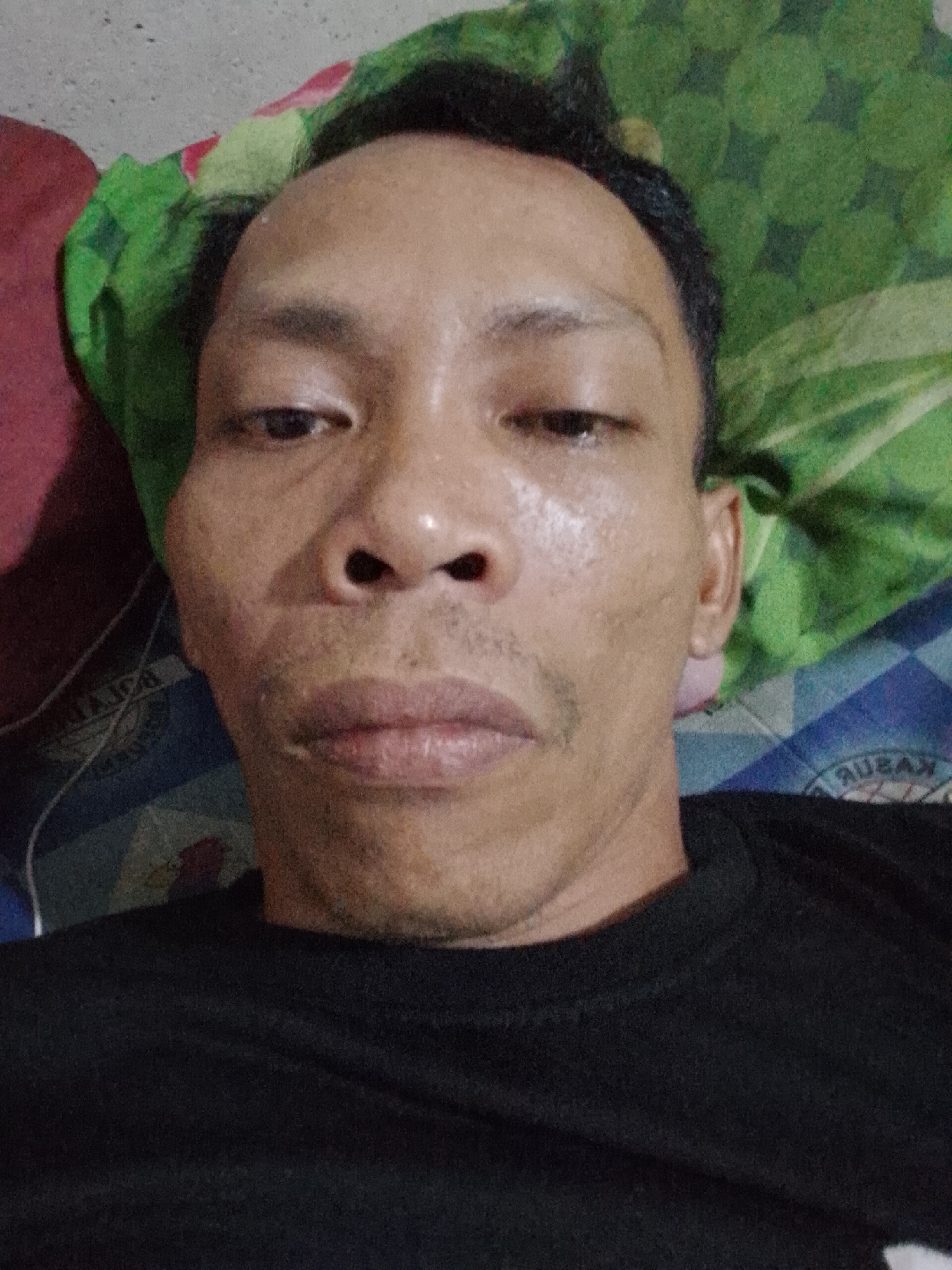 A selfie shot of a man, likely in his late 30s or early 40s, lying down. He has short, dark hair and a somewhat shiny face. The background shows a pillow with green and white patterns, possibly floral, and a bed with blue and white patterned sheets. The man is wearing a black t-shirt. The location is Bukit Ketok, Indonesia.