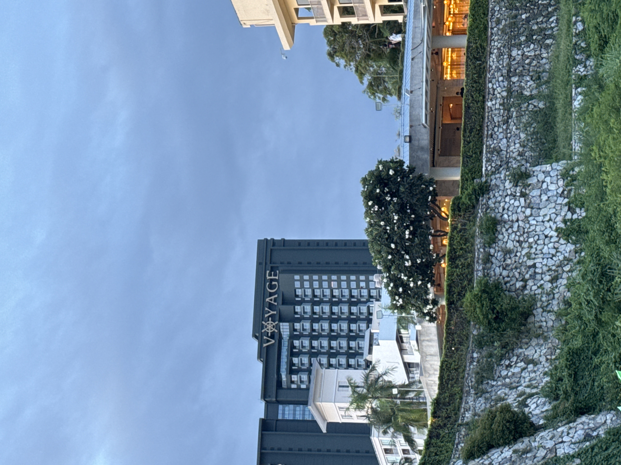 This image captures a section of a hotel in Pattaya City, Thailand, with a focus on its facade and the surrounding landscape. The prominent feature is a dark green, multi-story building with the word "V-YAGE" emblazoned in white lettering across its upper section. The building has a modern architectural style, characterized by numerous windows and balconies. To the left of this main building is a smaller, lighter-colored structure, and several palm trees are visible. In the foreground, a stone wall covered in lush green vegetation separates the hotel area from what appears to be a lower ground level, also densely vegetated. The sky above is overcast with a soft, diffused light, suggesting either early morning or late afternoon. Several individuals can be seen on balconies and within a covered outdoor area of the hotel, some appearing to be guests or staff. The overall impression is one of a coastal resort, likely at dusk or dawn, given the lighting conditions.