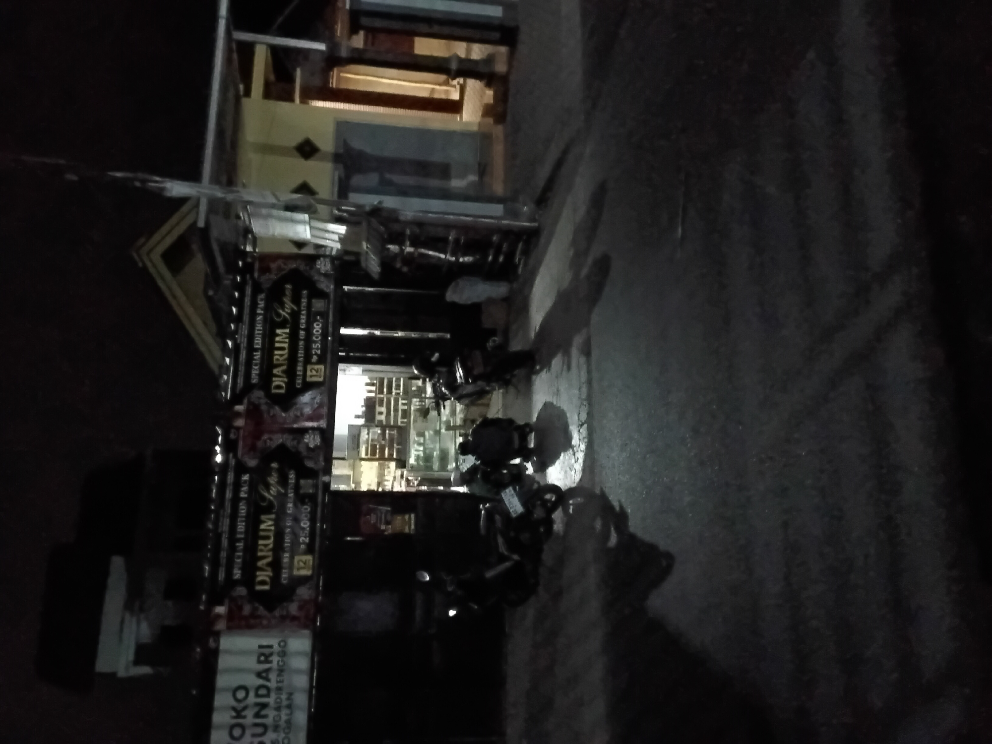The image depicts a nighttime street scene in Trenggalek, Indonesia, in front of a small storefront. The building appears to be a shop, possibly selling convenience goods, with shelves visible inside displaying various items. Two men are crouched on the ground outside the shop, one of them near a motorcycle. Another motorcycle is parked to the left of the frame. The storefront has signage, including advertisements for "DJARUM Super" cigarettes with "SPECIAL EDITION PACK" and pricing information. The lighting is dim, with the primary illumination coming from the shop's interior and some streetlights. The road surface is asphalt, and shadows are cast on it, indicating artificial light sources. The overall atmosphere is quiet and appears to be late at night.