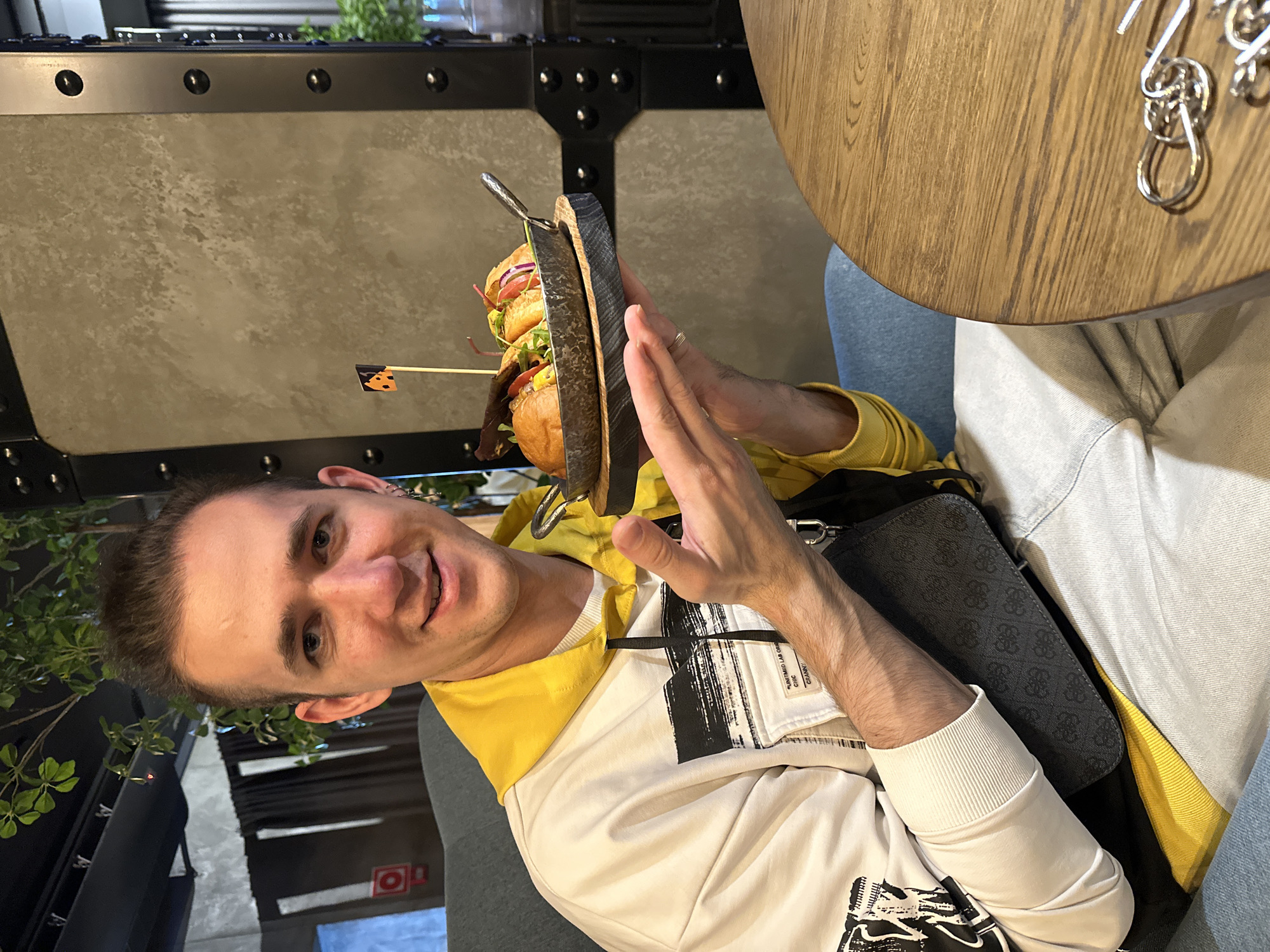 A person, identified as male, is seated at a table, holding a two-tier burger presentation in their hands. The burgers are stacked on a wooden board with a metal handle. The person is wearing a white and yellow sweatshirt. A purse is placed beside them. The location is identified as Saratov, Russia. The table is wooden and has a metal ring puzzle on its surface. The background features an industrial-style wall panel. A plant is visible in the far background.