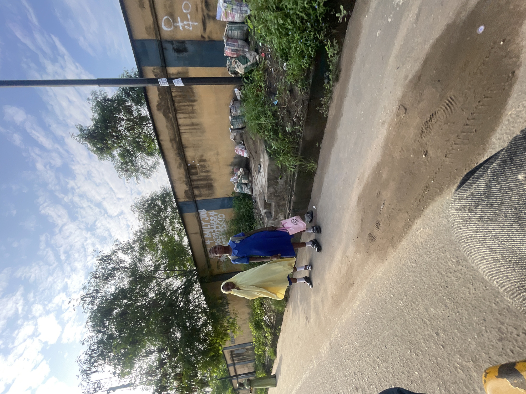 In Akowonjo, Nigeria, two young girls, one wearing a yellow hijab and the other a blue school uniform, stand on the side of a paved road. The girl in the hijab stands slightly behind the girl in the blue uniform, who is holding a pink tote bag. Behind them, a weathered, yellow and blue striped wall runs along the road, with overgrown vegetation in front of it. Several large, burlap sacks are piled along the base of the wall. A tall, dark lamppost stands near the wall, with stickers attached to it. In the background, trees with dense foliage are visible against a sky with scattered clouds. The foreground of the image shows a portion of the paved road and what appears to be the leg of a person, suggesting the image was taken from their perspective.