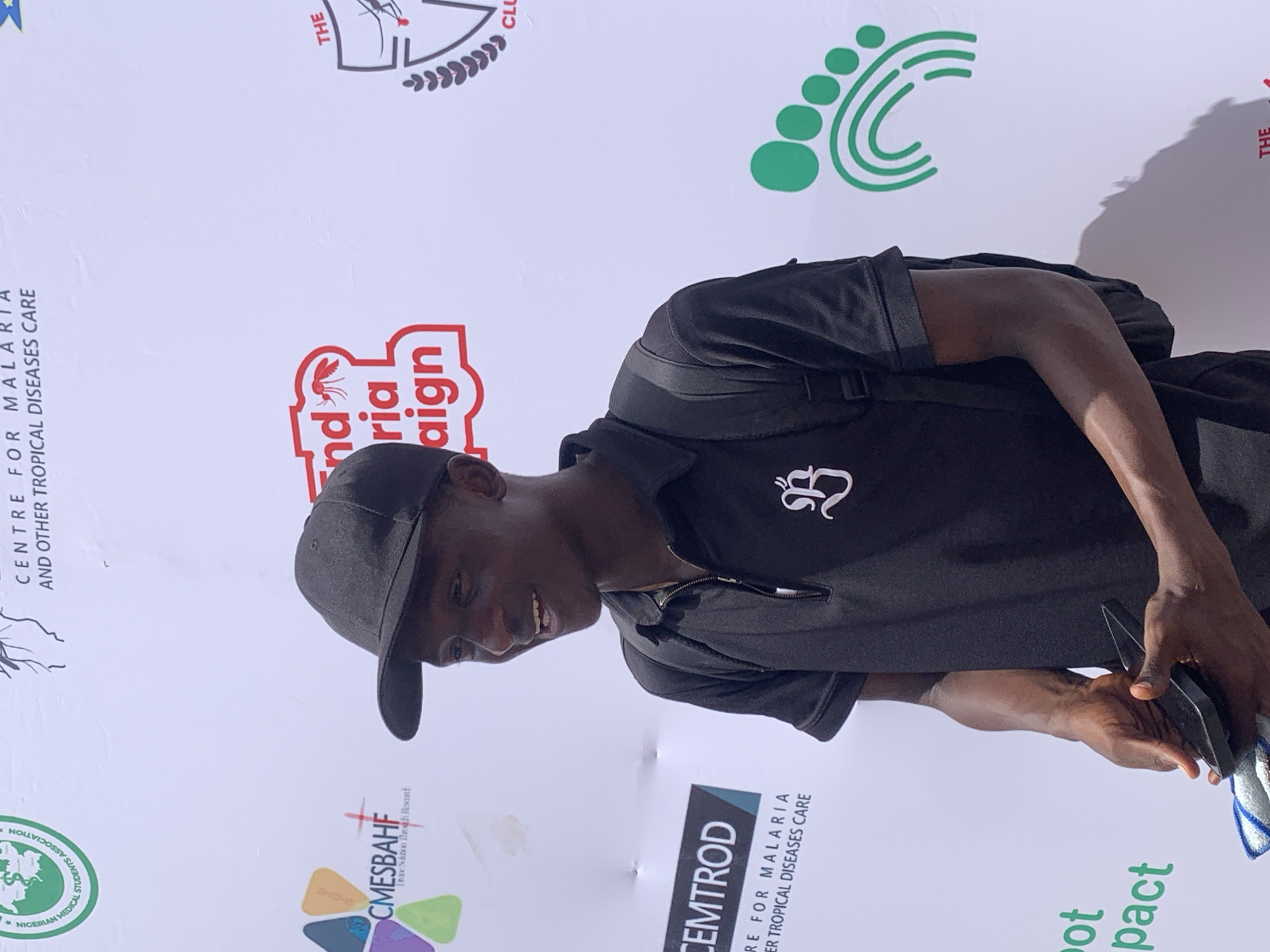 A young man of African descent, wearing a black cap and a black polo shirt with a white logo, is captured smiling while looking to his right. He has a black backpack on his shoulders and is holding a dark object, possibly a phone, in his hands. The setting is an event with a large white backdrop or banner displaying various logos and text, indicating an organizational or awareness campaign. The image appears to be rotated 90 degrees counter-clockwise.
Visible text and logos on the banner include:
* "CENTRE FOR MALARIA AND OTHER TROPICAL DISEASES CARE" (partially visible in multiple places).
* "NIGERIAN MEDICAL STUDENTS ASSOCIATION" with its logo.
* "CMESBAHF" with the tagline "Dare Sustain Though Research" (partially visible).
* "CEMTROD".
* A prominent red and white logo reading "nd ria aign," likely part of an "End Malaria Campaign."
* A green logo depicting a footprint with concentric circles.
* A logo featuring a mosquito and a shield, along with "THE CLU" (likely "THE CLUB").
* The words "ot pact" are also visible, possibly related to the green footprint logo.
The man's positive expression suggests he is engaged or enjoying the event. Given the recurring themes of malaria, tropical diseases, and student associations, this scene, located in Ilorin, Nigeria, likely depicts an educational outreach or awareness event focused on public health issues. The lighting suggests it is daytime.