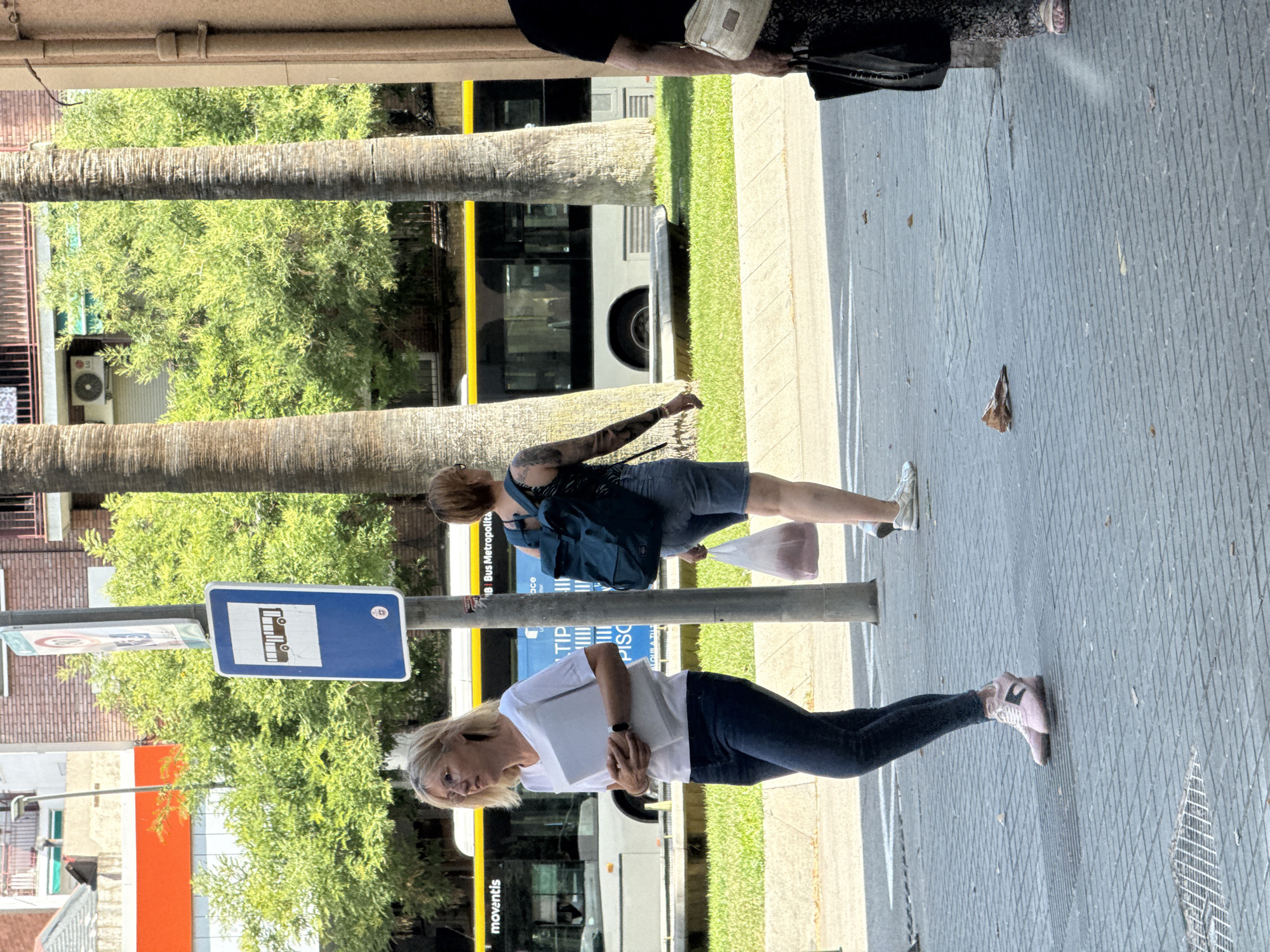 A street scene in L'Hospitalet de Llobregat, Spain, captures several individuals on a sunny day. In the foreground, a woman with blonde hair and glasses, wearing a white t-shirt and dark jeans, stands with a white folder in her arms. To her right and slightly behind, another woman with short brown hair and visible arm tattoos, wearing a dark patterned tank top and denim shorts, walks with a transparent plastic bag in her right hand. Further to the right, the lower half of another person is visible, wearing dark patterned trousers and a light-colored shoulder bag. A bus, identified by the "moventis" logo, is visible in the background, alongside a bus stop sign displaying a bus icon. Palm trees and leafy green trees provide a backdrop, with buildings and an air conditioning unit visible in the distance. The ground is paved with grey interlocking bricks.