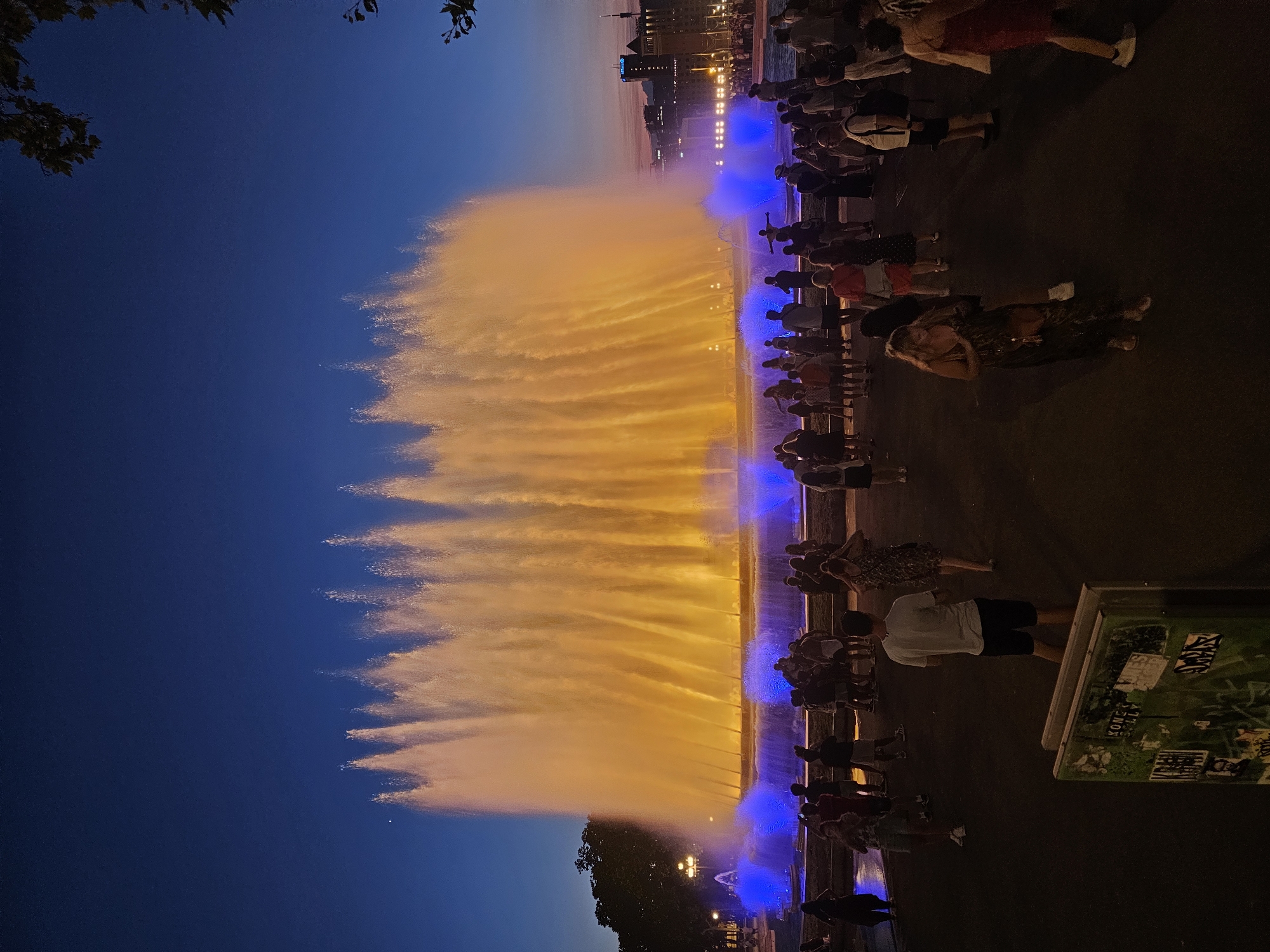 This media file depicts a nighttime view of the Magic Fountain of Montjuïc in Barcelona, Spain. The fountain is illuminated with vibrant orange and blue lights, creating a spectacular display of water jets against the dark sky. A crowd of people is gathered to watch the show, silhouetted against the colorful lights. The time of day is clearly evening or night, indicated by the dark sky and artificial lighting. The weather appears to be clear, allowing for the outdoor event. The architecture in the background suggests an urban setting, consistent with Barcelona. The overall atmosphere is one of awe and enjoyment, as people gather to witness the impressive water and light spectacle. There is a sign in the lower right corner with some text and graphics, but it is not clearly legible in the provided image.