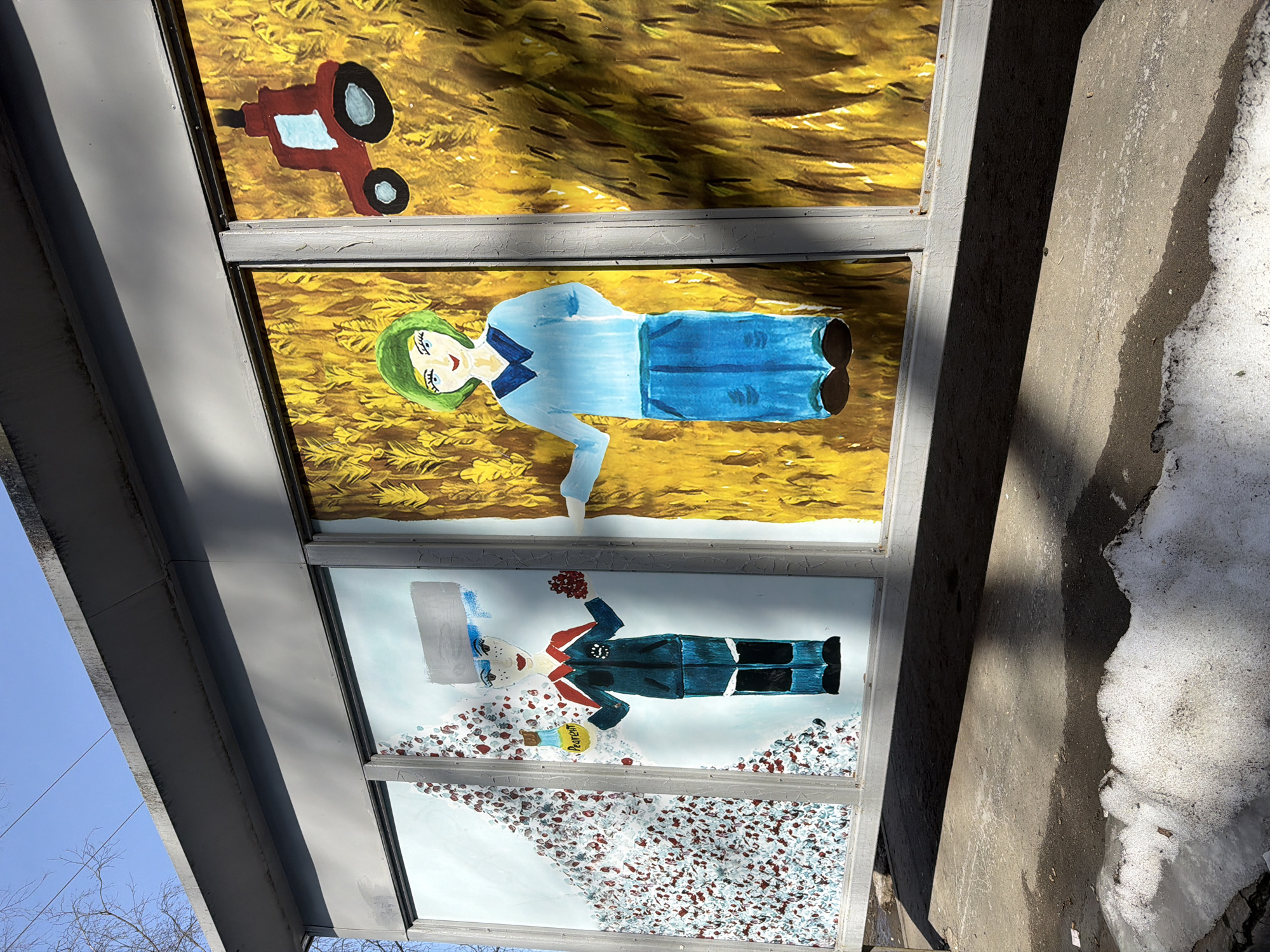 The image shows a bus stop in Berezniki, Russia, adorned with naive paintings. The top panel depicts a red tractor in a yellow field. The middle panel shows a woman with green hair wearing a light blue dress in the same field. The bottom panel features a figure in a dark blue military-style uniform with red accents, holding flowers and a container labeled "Perm," standing amidst a landscape of white and red scattered shapes, possibly representing snow and cranberries. A concrete curb with melting snow runs along the right edge of the image. The sky is visible in the top left corner.