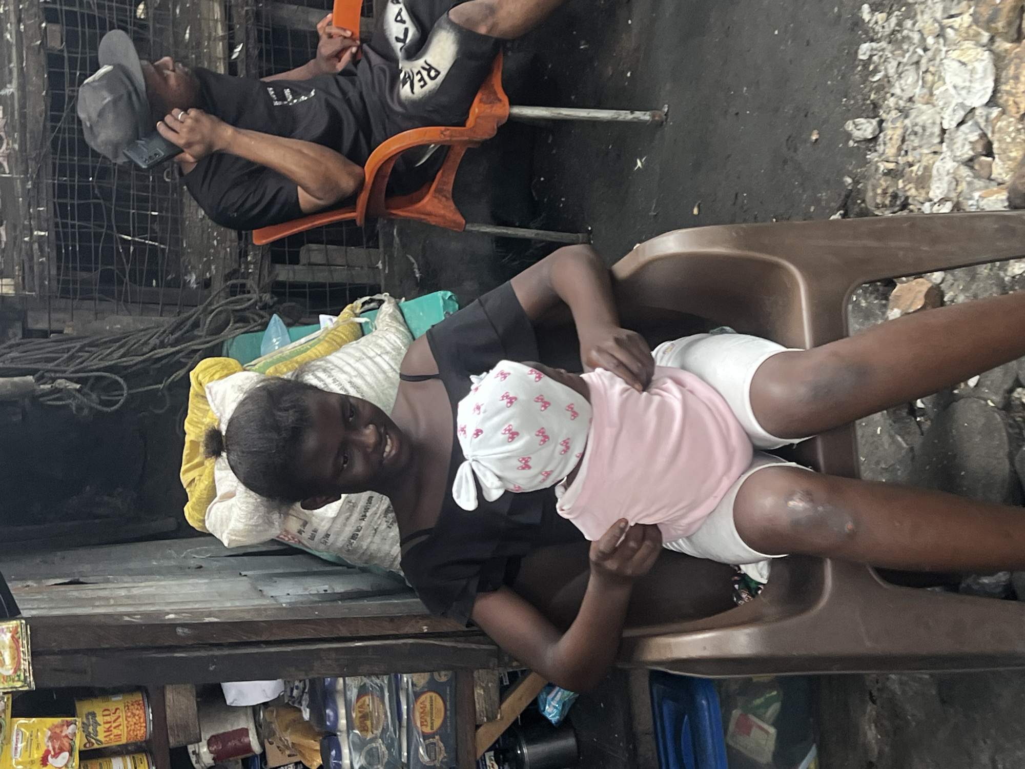 The image captures a candid scene in Nkpor, Nigeria. In the foreground, a young woman with dark skin and dark hair pulled into a bun is seated in a brown plastic chair. She is holding a baby dressed in a pink outfit and a white cap with pink bow patterns. The woman is looking down at the baby and smiling. Her attire includes a black off-the-shoulder top and white shorts. Her legs show some scraped or bruised areas on the knees.
To the right, an orange plastic chair is occupied by a man wearing a black t-shirt and a gray baseball cap, talking on a mobile phone. He is looking to the right.
The background is cluttered with various objects, suggesting a market or outdoor stall setting. There are shelves stocked with canned goods and packaged items. Large sacks, one yellow and others white, are piled up behind the woman. The ground is dirt and covered with small rocks and debris. The overall environment appears rustic and somewhat unkempt, typical of many urban street scenes in Nigeria. The lighting suggests it is daytime.