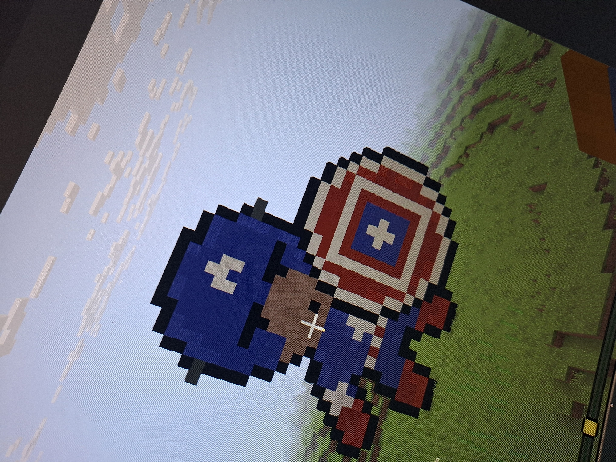 The image shows a pixelated representation of Captain America, rendered in a style reminiscent of Minecraft. The character is depicted from the chest up, holding a shield. The background appears to be a sky with pixelated clouds and a green landscape with some brown patches, suggesting a game environment. There are no discernible features that would allow for the identification of a specific location in Varna, Bulgaria, or any other real-world place. The image is a digital creation.