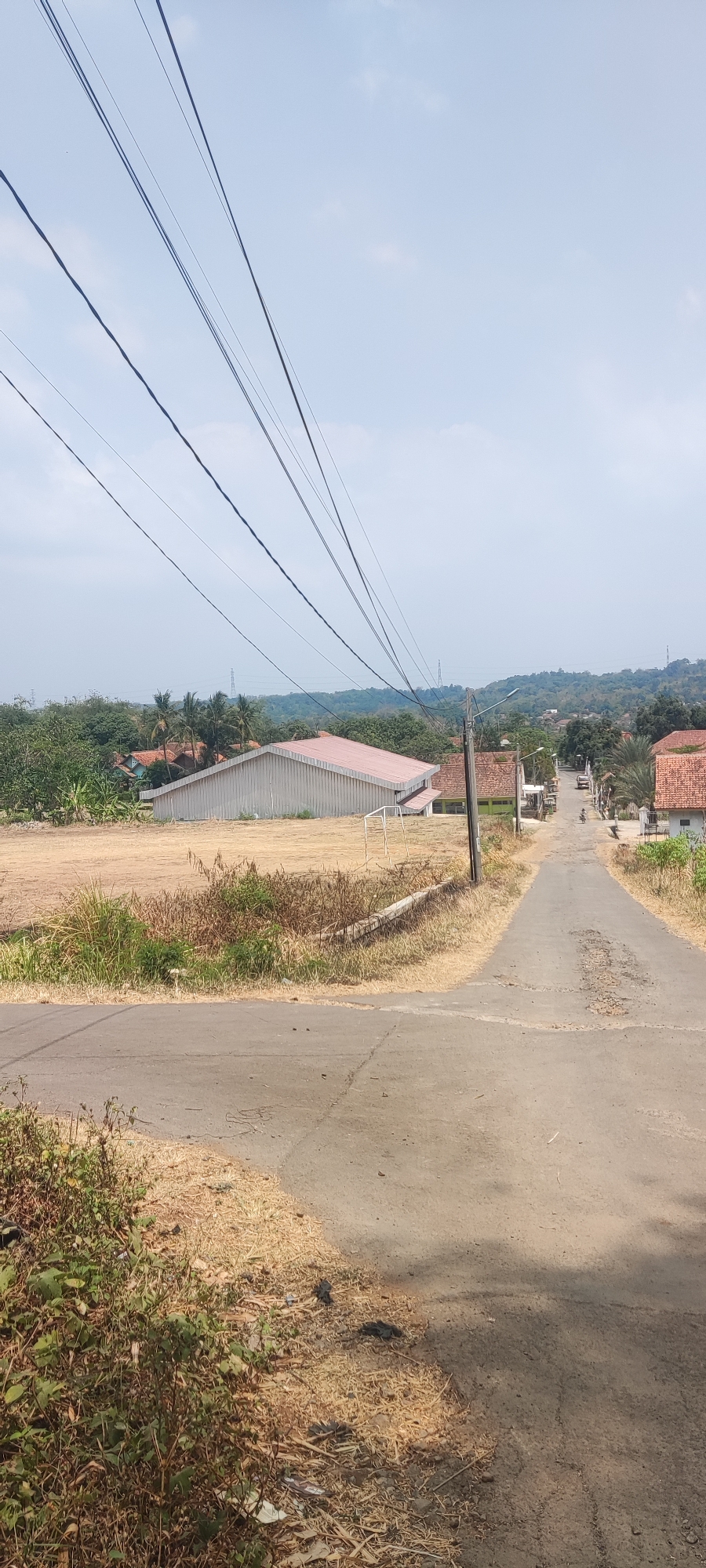 The image depicts a rural street scene in Panongan Lor, Indonesia. The foreground shows a cracked asphalt road with dry, sparse vegetation along its edges. To the left, a large, light-colored building with a reddish-brown roof sits on a dry, open field, adjacent to a soccer goal. In the background, a winding road leads uphill, with houses and trees lining its sides. Power lines stretch across the sky, and in the distance, rolling hills are visible. The sky is partly cloudy and appears bright, suggesting daylight. A single person on a motorcycle is visible on the road in the mid-ground.
