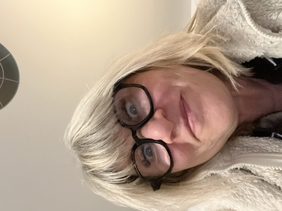 A woman with blonde hair and black-rimmed glasses looks into the camera. The image is rotated, with the top of the frame oriented to the left. She has fair skin and is wearing a fluffy, light-colored jacket. A circular object is visible in the upper left corner. The woman's expression is slightly quizzical, and her eyes are visible through her glasses. The background appears to be a plain, light-colored wall.