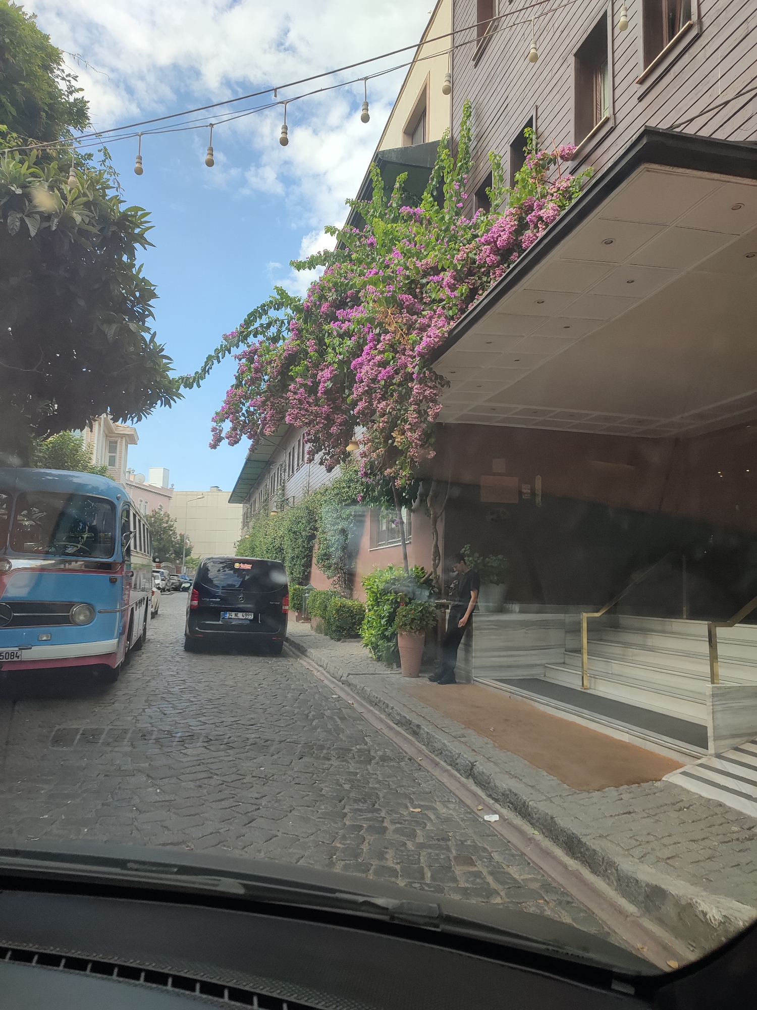 The image depicts a street scene in Istanbul, Turkey, likely in a historic district given the cobblestone road and architecture. The view is from inside a vehicle, looking out through the windshield. A blue and red retro-style bus is parked on the left side of the narrow, cobblestone street. To its right, a black Mercedes-Benz Vito van is also parked, displaying a Turkish license plate.
The buildings lining the street have a rustic charm. The building on the right is a multi-story structure with dark wood siding and windows. A vibrant cascade of pink bougainvillea flowers spills over the edge of an overhang from the upper stories, creating a striking visual contrast against the dark wood and the blue sky. A man dressed in black stands near the entrance of this building, leaning against a planter.
The sky is a clear blue with scattered white clouds. String lights with bare bulbs are visible overhead, suspended across the street, suggesting a decorative or functional lighting setup for the area, possibly for an evening ambiance. The overall impression is a charming, somewhat congested urban street with a blend of historical and modern elements.