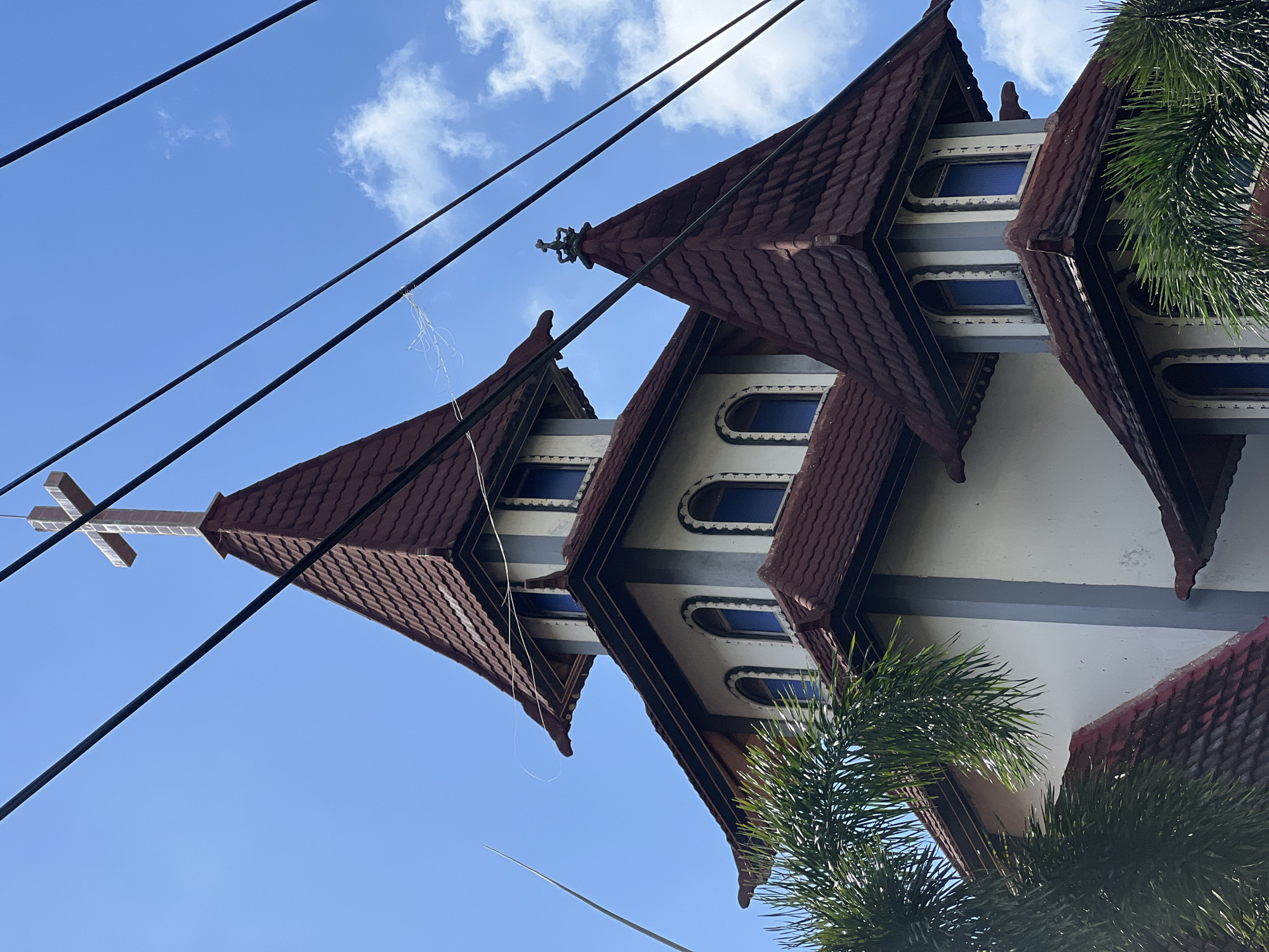 An upward, close-up, low-angle shot captures the upper section of a church with distinctive tiered, conical red-tiled roofs, set against a bright blue sky with scattered white clouds. The church building features a white facade with decorative grey trim around arched windows that contain blue glass. A prominent cross adorns the apex of the tallest spire. Multiple black electrical wires crisscross the frame diagonally, obscuring parts of the sky and the upper structure of the church. To the left and right, the fronds of green palm trees intrude into the frame, adding a tropical element to the scene. The image appears to have been taken in a built-up area, given the presence of the electrical wires. This church is located in North Kuta, Indonesia.