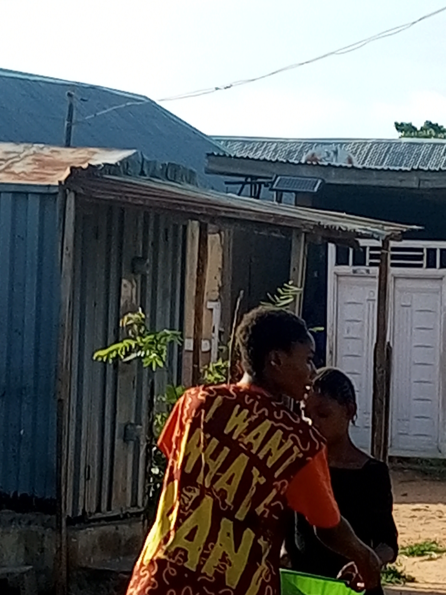 In Tempure, Nigeria, two young individuals are partially visible outdoors. The person in the foreground, wearing a red shirt with yellow lettering that appears to read "I WANT WHAT I WANT," is facing to the right. Behind them, another individual in a black shirt is also facing to the right. They are situated in front of a weathered structure with corrugated metal siding and exposed wooden supports. In the background, a building with white doors and a corrugated metal roof is visible, with trees and a power line overhead.