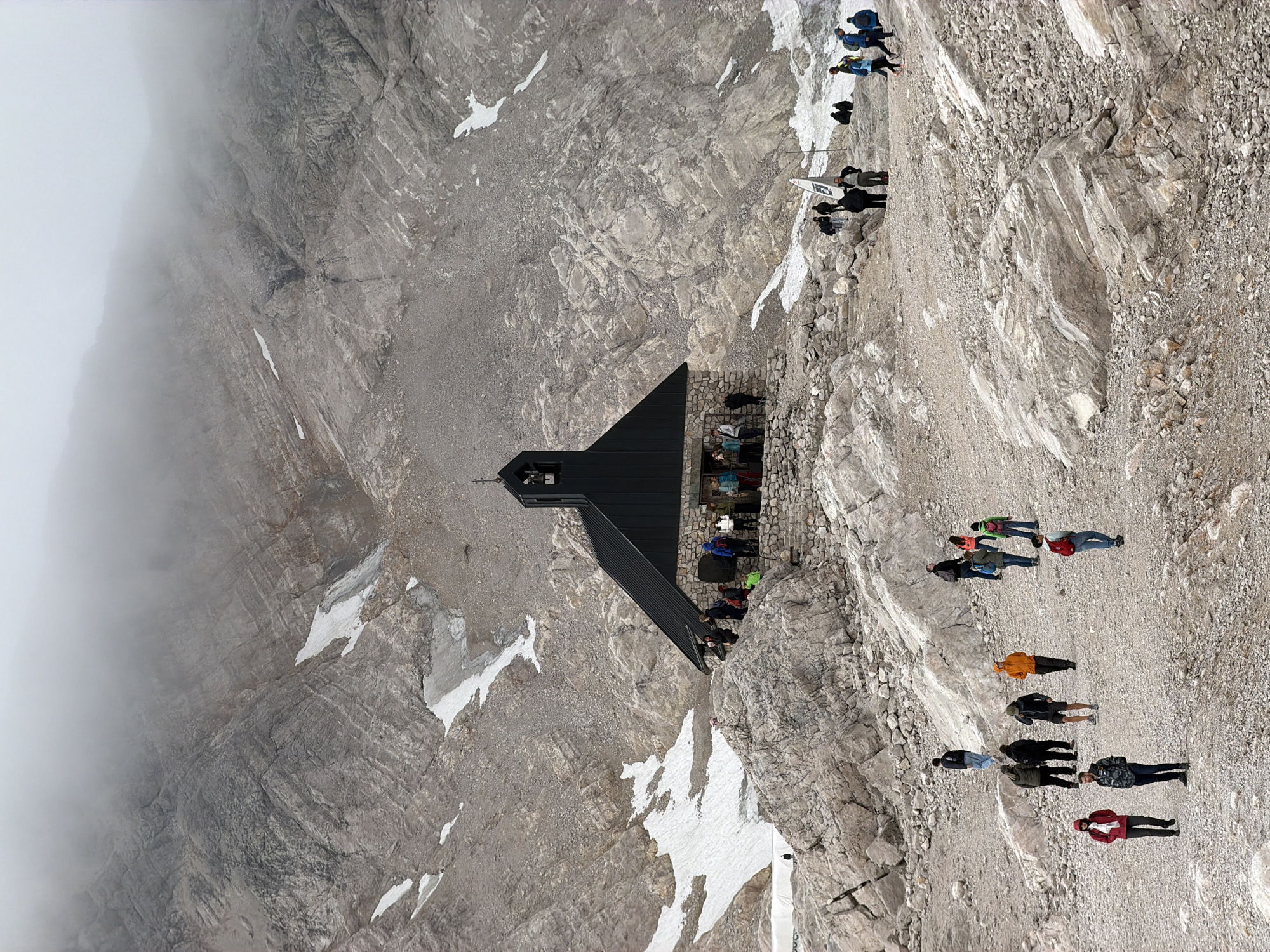 The image depicts the Agnes-Bernauer Chapel on the Zugspitze, the highest peak in Germany, located near Garmisch-Partenkirchen. The chapel, a dark, angular structure with a stone base, sits on a rocky, mountainous terrain dotted with patches of snow. A group of tourists are seen gathered around the chapel and walking on the gravelly paths in front of it. The background is dominated by rugged, grey rock faces partially obscured by a thick layer of fog or clouds, creating a dramatic and somewhat ethereal atmosphere.