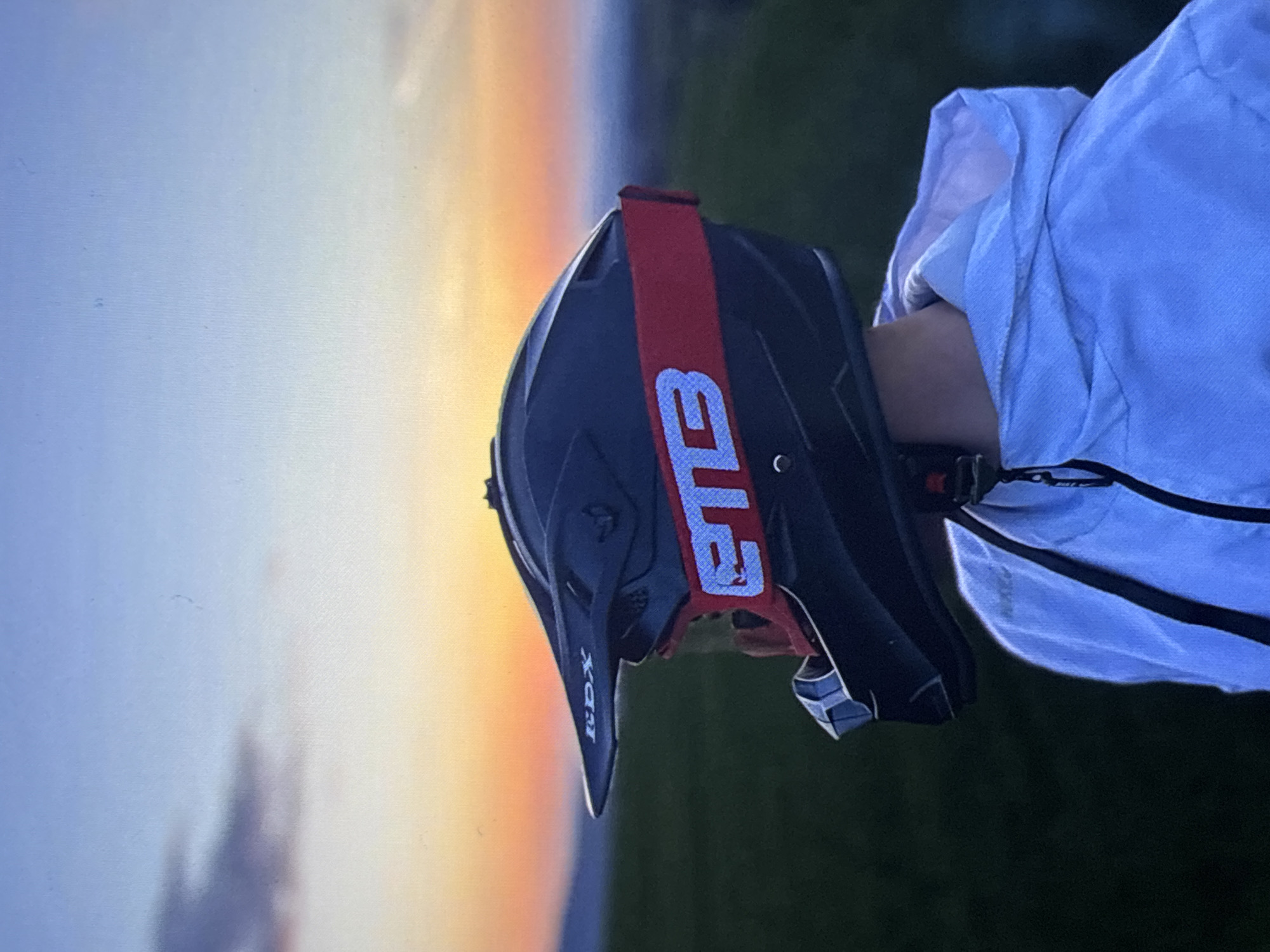 A dirt bike helmet with a red strap sits on a person's arm, with a sunset and green fields in the background. The helmet is black and has the brand name "RDX" on the front. The sunset appears to be setting over an area of green grass. Location: Unknown, Unknown.