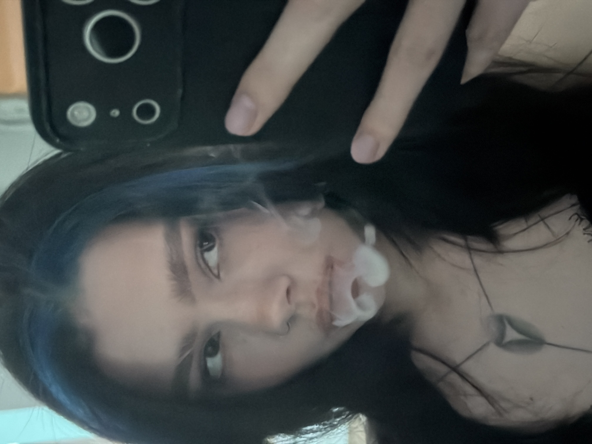 The image is a mirror selfie capturing a young person, likely female, with dark hair featuring blue streaks. She looks directly into the camera/mirror with a neutral expression. A small ring piercing is visible in her nose. Wisps of white smoke, forming partial rings, are exhaled from her mouth, indicating an act of vaping or similar. There is a reddish discoloration around her lips.
The subject is holding a black smartphone, partially visible at the top, with her fingers wrapped around the phone case. The case shows distinct camera lenses and sensors. Around her neck, a necklace with a dark pendant adorned with a lighter, abstract triangular design is visible. The background is mostly obscured by her hair and clothing, suggesting an indoor setting with diffused lighting. The activity takes place in Khimki, Russia. No visible text is present in the image.