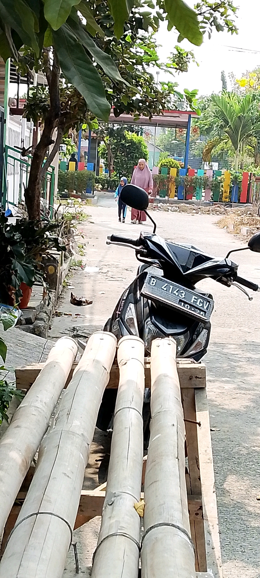 In the city of Satriamekar, Indonesia, a scene unfolds on an asphalt pathway lined with vegetation. In the foreground, a black motorcycle is parked, its license plate reading "B 4143 FGV 10-26". Resting in front of the motorcycle is a structure made of four large bamboo logs, laid horizontally on wooden planks. To the left of the path, a leafy tree shades potted plants. Further down the path, a woman in a pink hijab and dress walks alongside a child in a blue jacket. Behind them, a colorful fence encloses an area with palm trees. The environment suggests a sunny day, possibly in a residential or public park area.