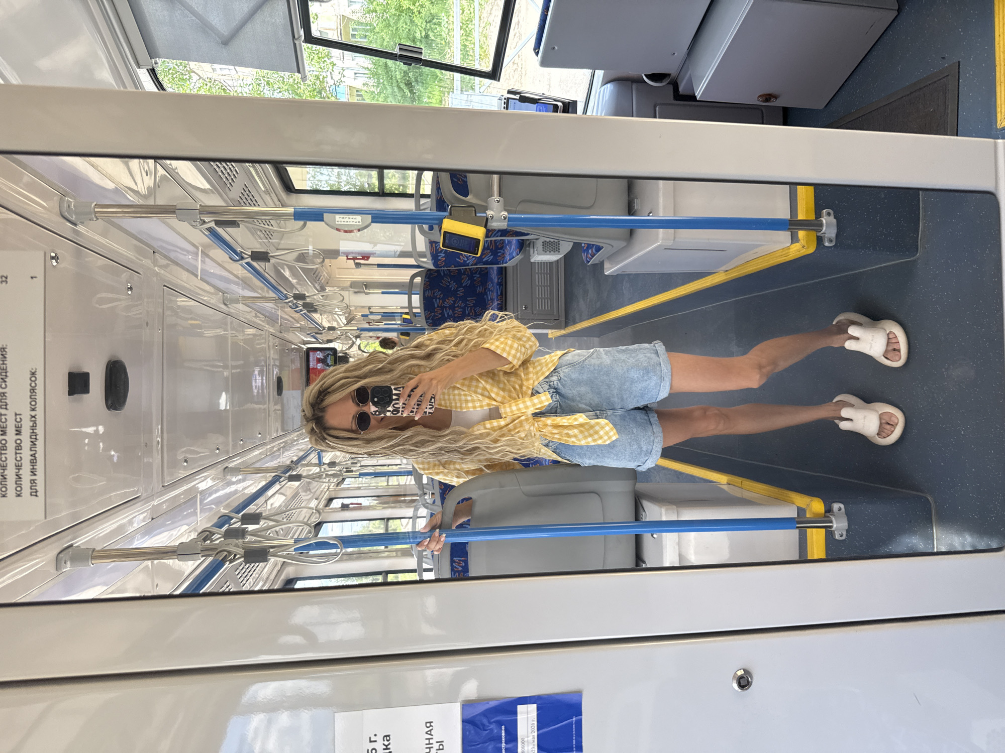 The image shows a woman standing inside what appears to be a bus or trolleybus. She is fair-skinned with long blonde curly hair, wearing round sunglasses, a yellow gingham shirt tied at the waist, and blue denim shorts. Her feet are clad in white slide sandals. She is holding a phone in front of her face, likely taking a selfie.
The interior of the bus or trolleybus is visible in the background. There are blue and yellow handrails, gray seating, and signs in Russian. Through the front window, green trees and buildings are visible.
Signs on the bus have some OCR text.
