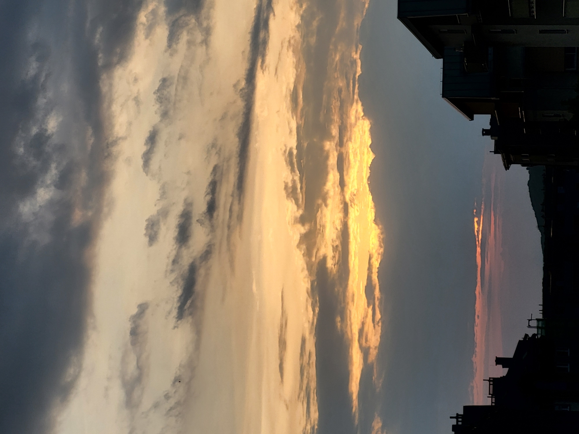 The image depicts a sky at dusk in an urban setting. The sky is filled with clouds, with some areas illuminated in golden light, likely from the setting sun. The right side of the frame shows silhouettes of buildings and structures against the fading light of the sky. There are no people visible in the image. The location is indicated as a city named Unknown, Unknown.
