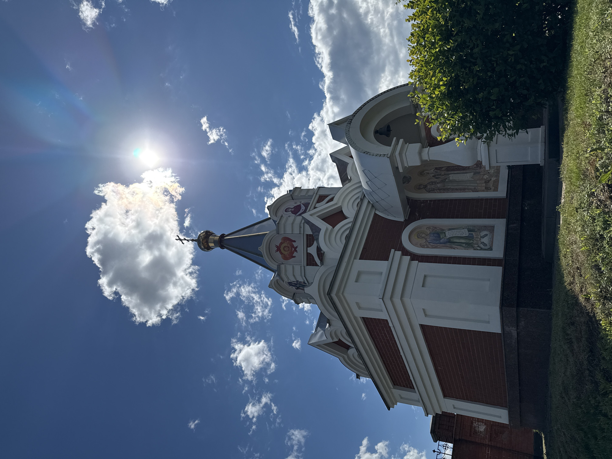 This is a low-angle, full shot of a small, ornate Orthodox chapel under a bright, partially cloudy sky. The sun is high and slightly to the right, creating lens flare and highlighting some of the clouds. The chapel is constructed with red brick and white trim, featuring a dark-colored, multi-tiered roof topped with a golden dome and cross. There are decorative religious murals on the exterior walls, depicting figures in traditional religious attire. The chapel has an arched entrance supported by white columns, and a bell is visible inside the archway. Lush green bushes and grass surround the base of the chapel, and a brick wall with a decorative metal fence is visible to the left. The location is identified as the Chapel of the Archangel Michael in Izhevsk, Russia. There are no people visible in the image.