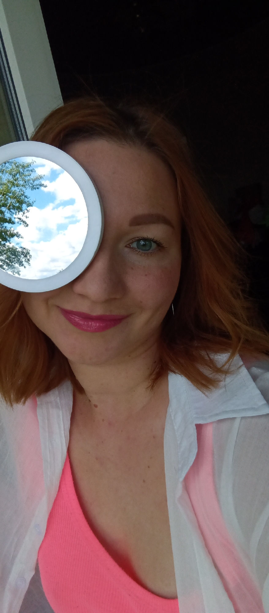 A Caucasian woman with red hair is depicted in a close-up shot. She is wearing a pink top underneath a white shirt and is smiling. She is holding a round mirror over her left eye. The mirror reflects a tree and a blue sky with clouds. The background behind the woman appears dark. The location is the city of Unknown, Unknown.