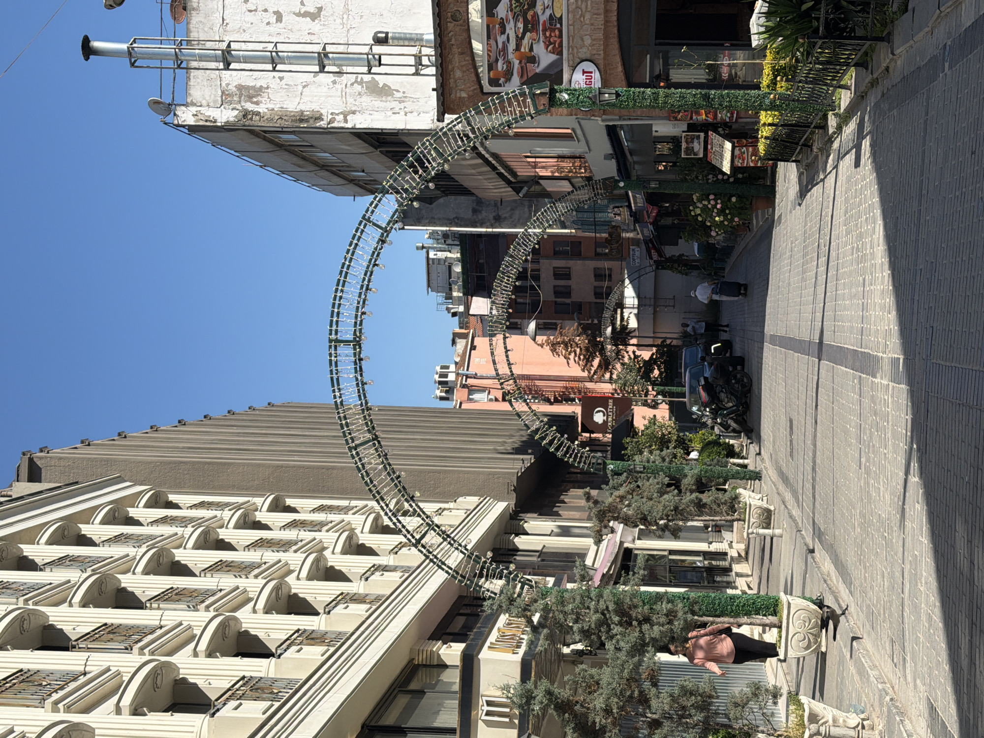 This image captures a street scene in Istanbul, Turkey, likely within a commercial or tourist area, given the presence of shops, restaurants, and the overall architecture. The day is clear and sunny, casting distinct shadows on the cobblestone street.
Dominating the foreground is a decorative, arched metal structure adorned with small lights, spanning across the street. On the left side of the street, a modern, multi-story building with cream-colored facade and numerous balconies with dark railings is prominently featured. Adjacent to it are trees in large planters.
On the right side of the street, buildings with a more traditional brick facade are visible, housing businesses with awnings and signage. One sign displays "DOUBLETREE BY HILTON." Further down the street, more buildings of varying architectural styles are apparent, including a tall, older building with a white, weathered exterior.
Several people are present in the scene. On the left sidewalk, a woman in a pink shirt and dark pants walks away from the camera. Further down the street, a person in white is seen pulling a wheeled suitcase. A few other figures are sparsely distributed along the street.
Objects visible include parked motorcycles, cars, street lamps, and various shop displays and advertisements. The environment is an urban street with a clear blue sky overhead.