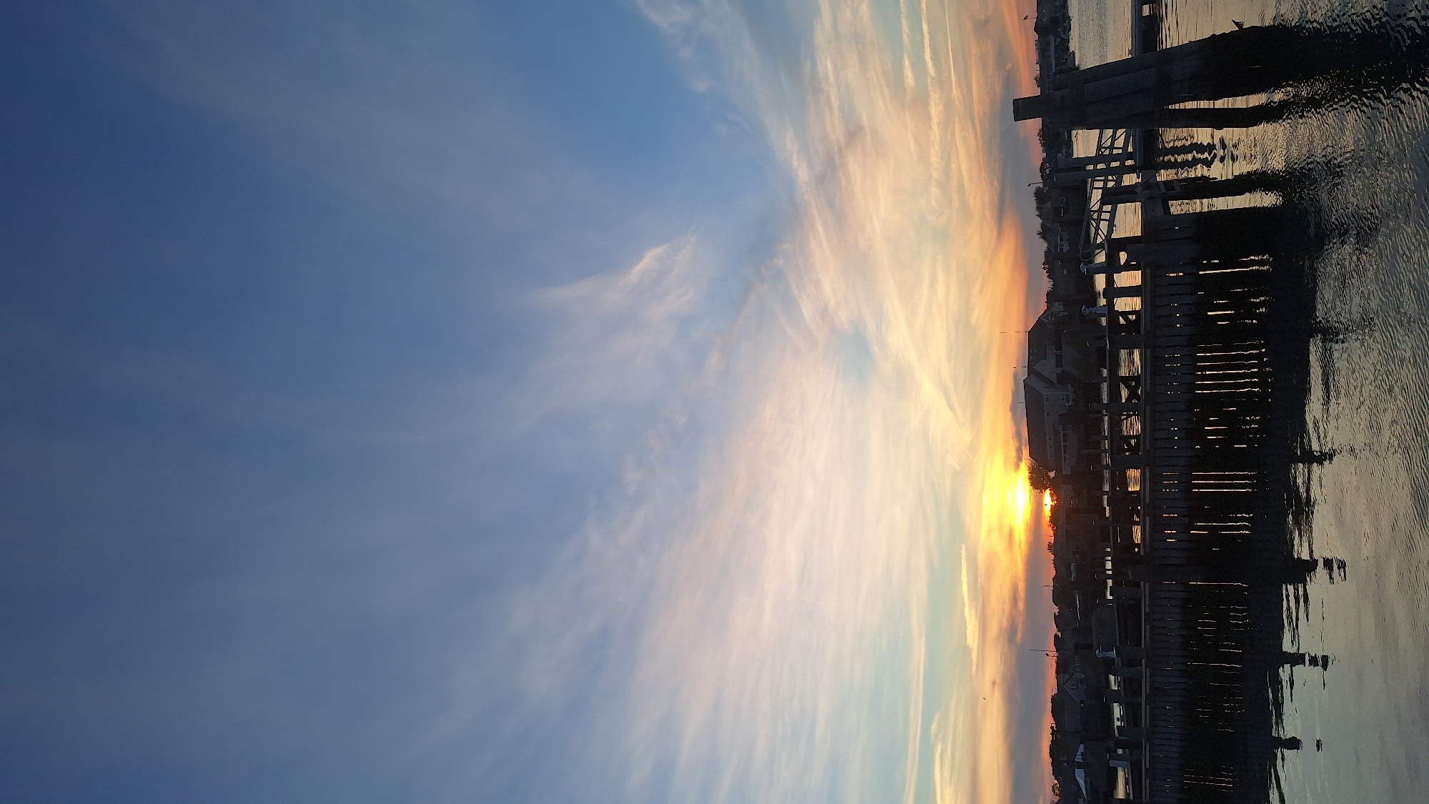 A vertical, medium shot captures a serene sunset over the water in Nantucket, Massachusetts. The sky is a gradient of deep blue at the top, transitioning to softer blues and pinks as it approaches the horizon, with wispy clouds streaked across. The sun, a bright orange orb, is just beginning to dip below the horizon, casting a warm glow on the scene. In the foreground, a weathered wooden dock juts out into the calm water, its pilings casting dark reflections. Buildings with pitched roofs are visible in the distance, silhouetted against the vibrant sunset. The water's surface is rippled, reflecting the colors of the sky and the dock.