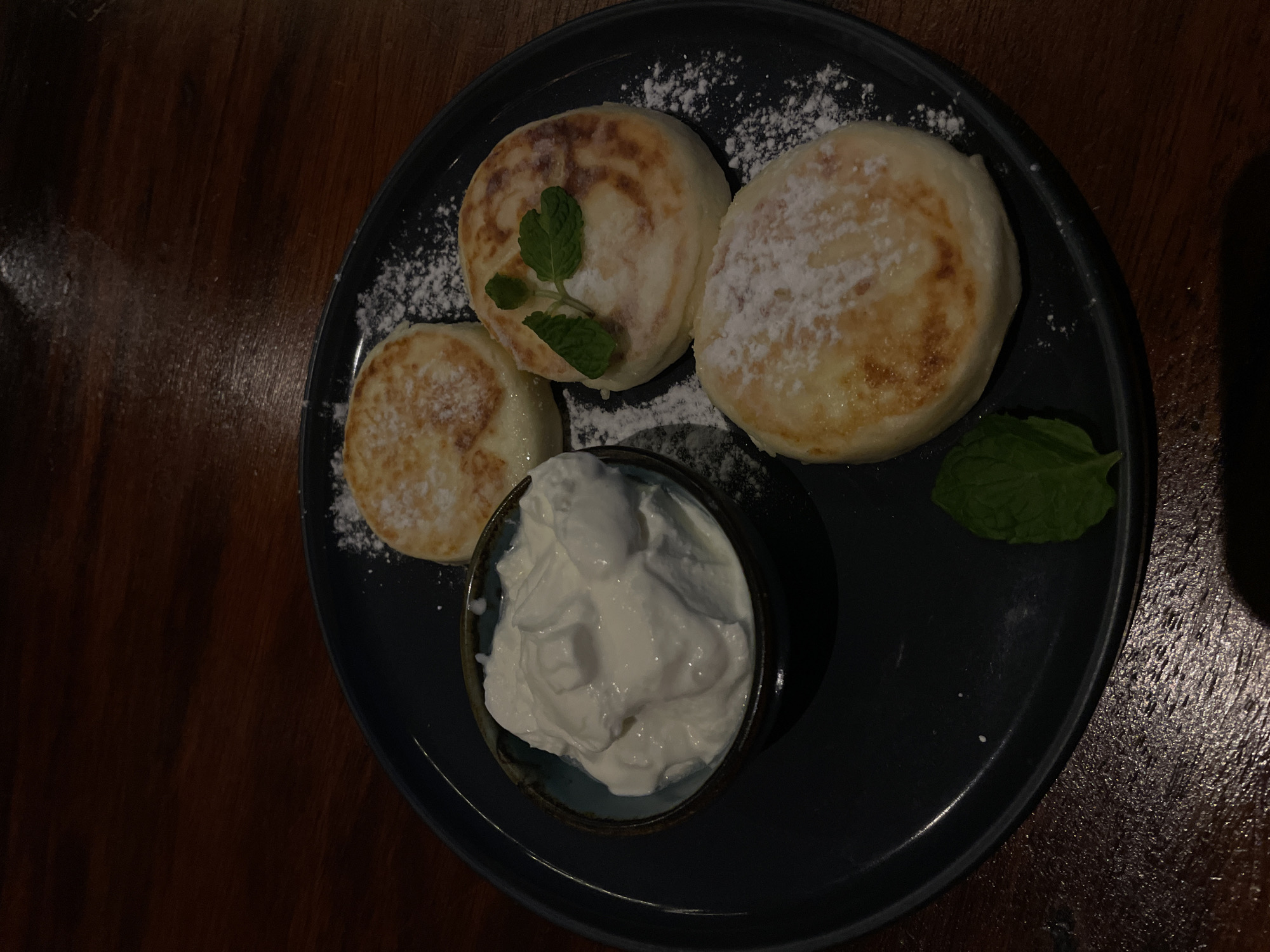 A dark-rimmed plate holds three round, golden-brown syrniki, a type of Russian cheese pancake, dusted with powdered sugar. One syrnik is garnished with two mint leaves. To the left, a small, dark blue bowl contains a generous serving of thick, white sour cream or yogurt. The plate rests on a dark, polished wooden surface, likely a table, with a dark background behind it. The scene is set in a dimly lit restaurant in North Kuta, Indonesia.