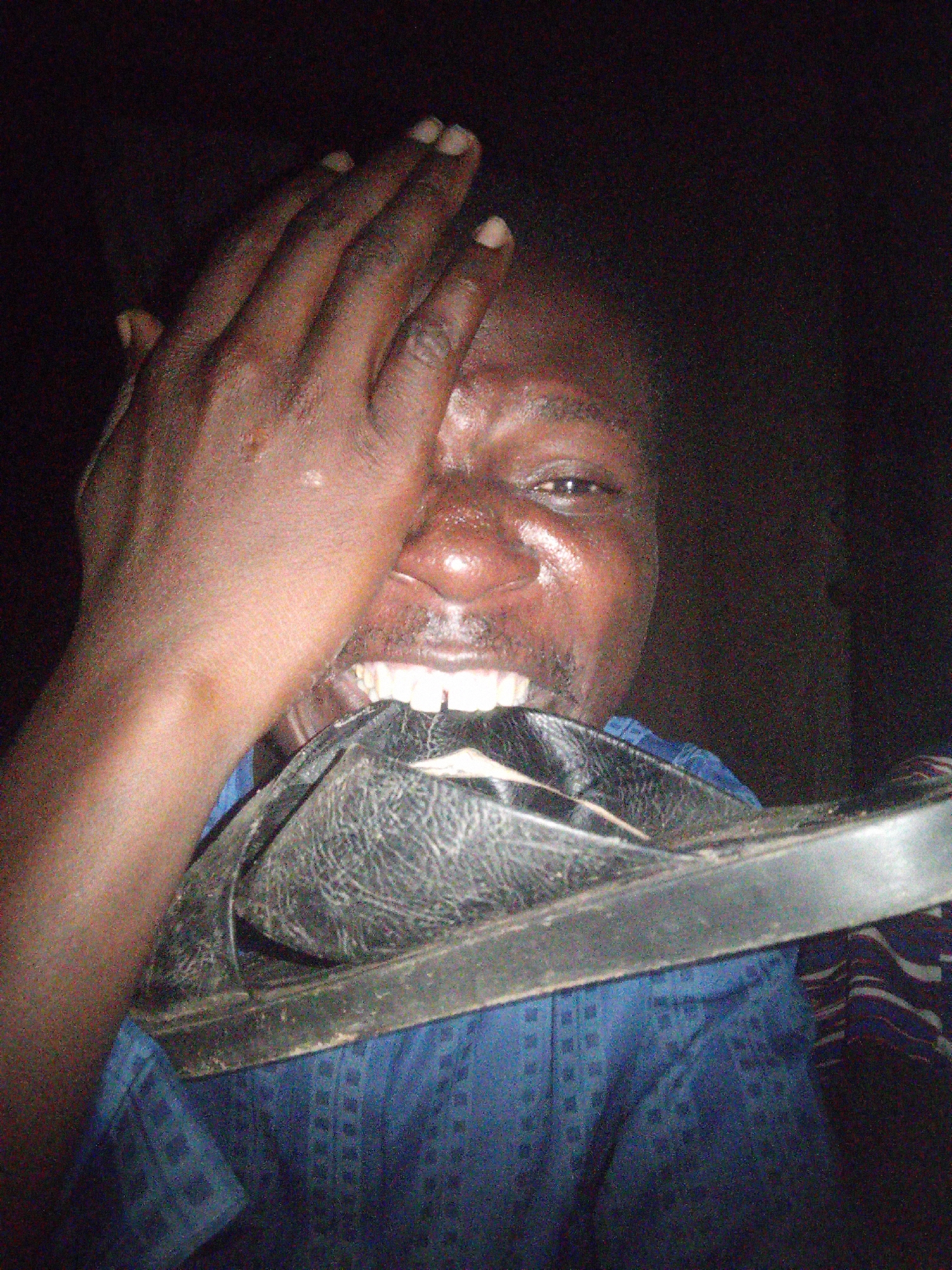 The image shows a close-up of a dark-skinned person, possibly a man, covering their face with their right hand. The person is wearing a blue patterned garment. A black sandal is positioned in front of the person's mouth and chin. The background is dark, suggesting an indoor or nighttime setting. The image quality is poor, with significant noise and low resolution. There is no identifiable information within the image to determine its specific location in Gombe, Nigeria, or any broader environmental context.
