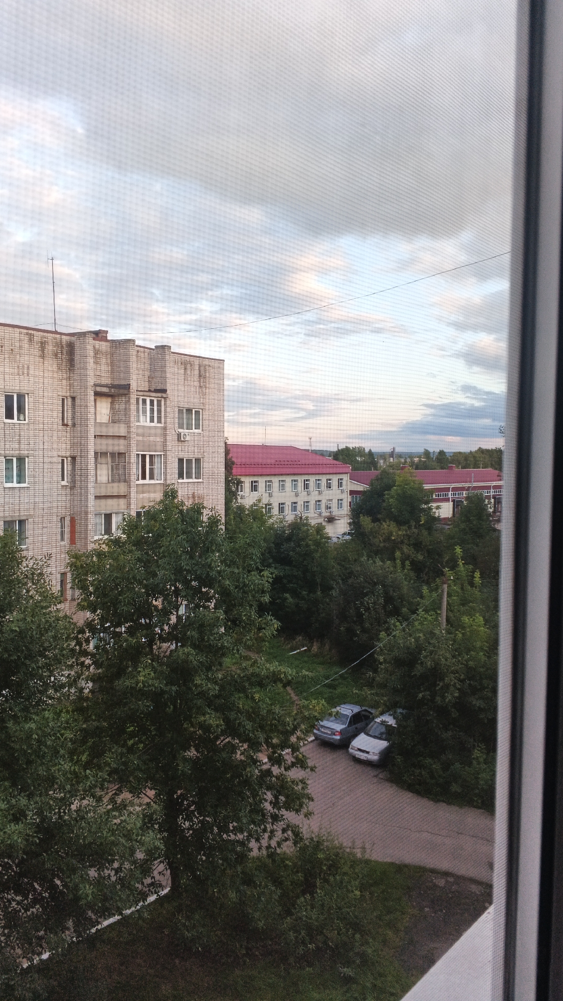 The image depicts a view from a window overlooking a residential area in Izhevsk, Russia. A multi-story apartment building constructed from light-colored bricks dominates the left side of the frame. The building features numerous windows, some with white frames and others with visible air conditioning units. Adjacent to this building, a longer, lighter-colored structure with a bright red roof is visible. This building also has many windows and air conditioning units. In the foreground, lush green trees and foliage obscure part of the view. A paved area with two cars parked is visible in the middle ground, surrounded by dense greenery. The sky above is overcast with a mix of white and gray clouds, suggesting a potentially cool or rainy day. A power line stretches across the sky. The image appears to be taken from a residential apartment, looking out onto a typical urban landscape.