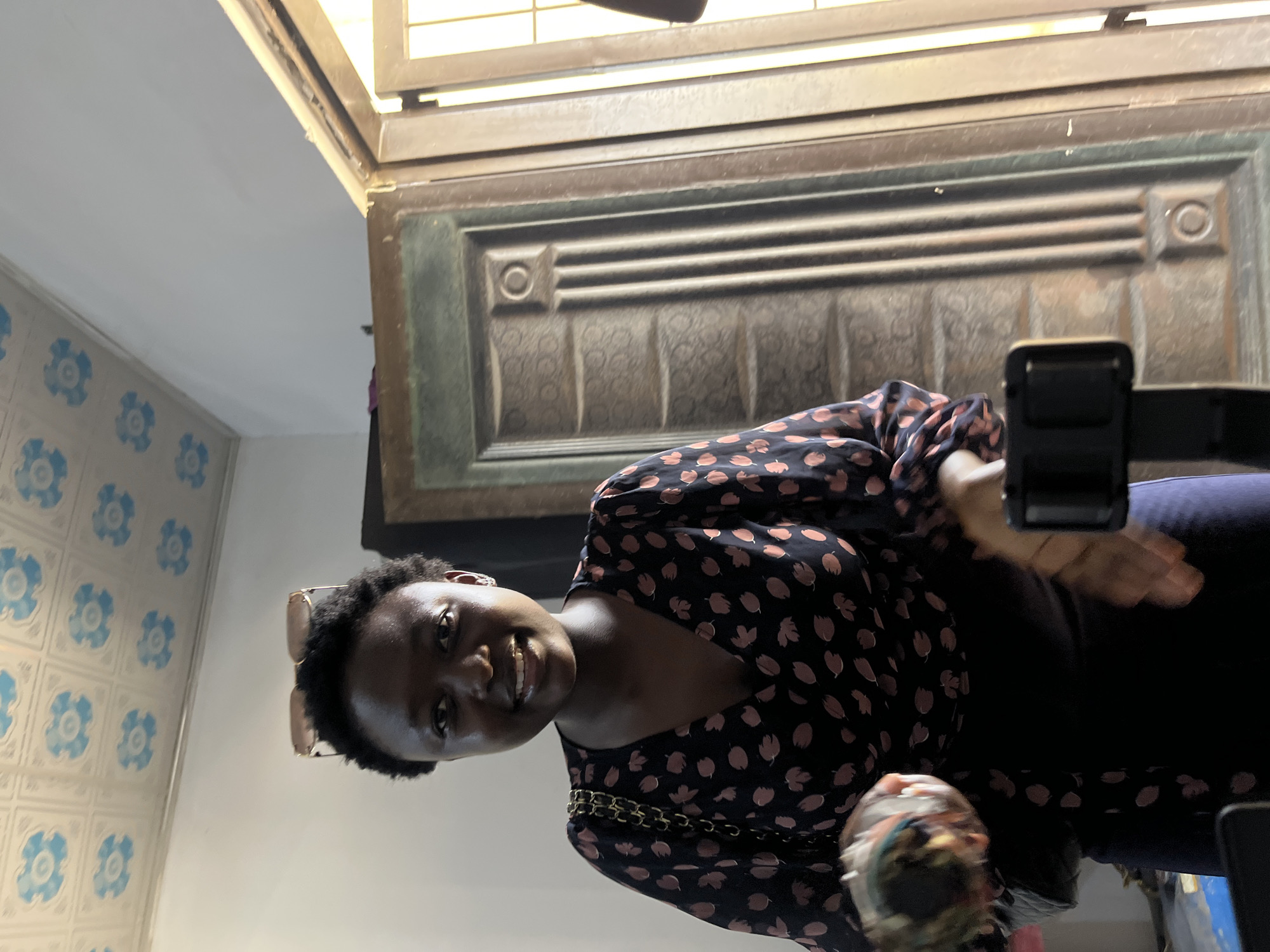 This media file depicts a young woman in a room with patterned wallpaper and a large, textured wooden door or panel in the background. The woman is smiling and looking towards the camera. She is wearing a dark, long-sleeved top with a floral or leaf-like pattern of pinkish-orange. A gold chain is visible over her shoulder, possibly part of a purse. She is also wearing sunglasses perched on her head.
The scene appears to be indoors, with a light source coming from above, casting shadows. The wallpaper on the left side of the image has a repeating pattern of blue floral motifs. The large wooden panel behind her has a grid-like design with horizontal grooves and decorative square insets. The overall lighting suggests it could be daytime. The image is taken from a low angle, looking upwards. The woman's expression is happy and engaging. The setting, a room in Gura Top, Nigeria, is characterized by these distinctive decorative elements. There is no visible text in the image.