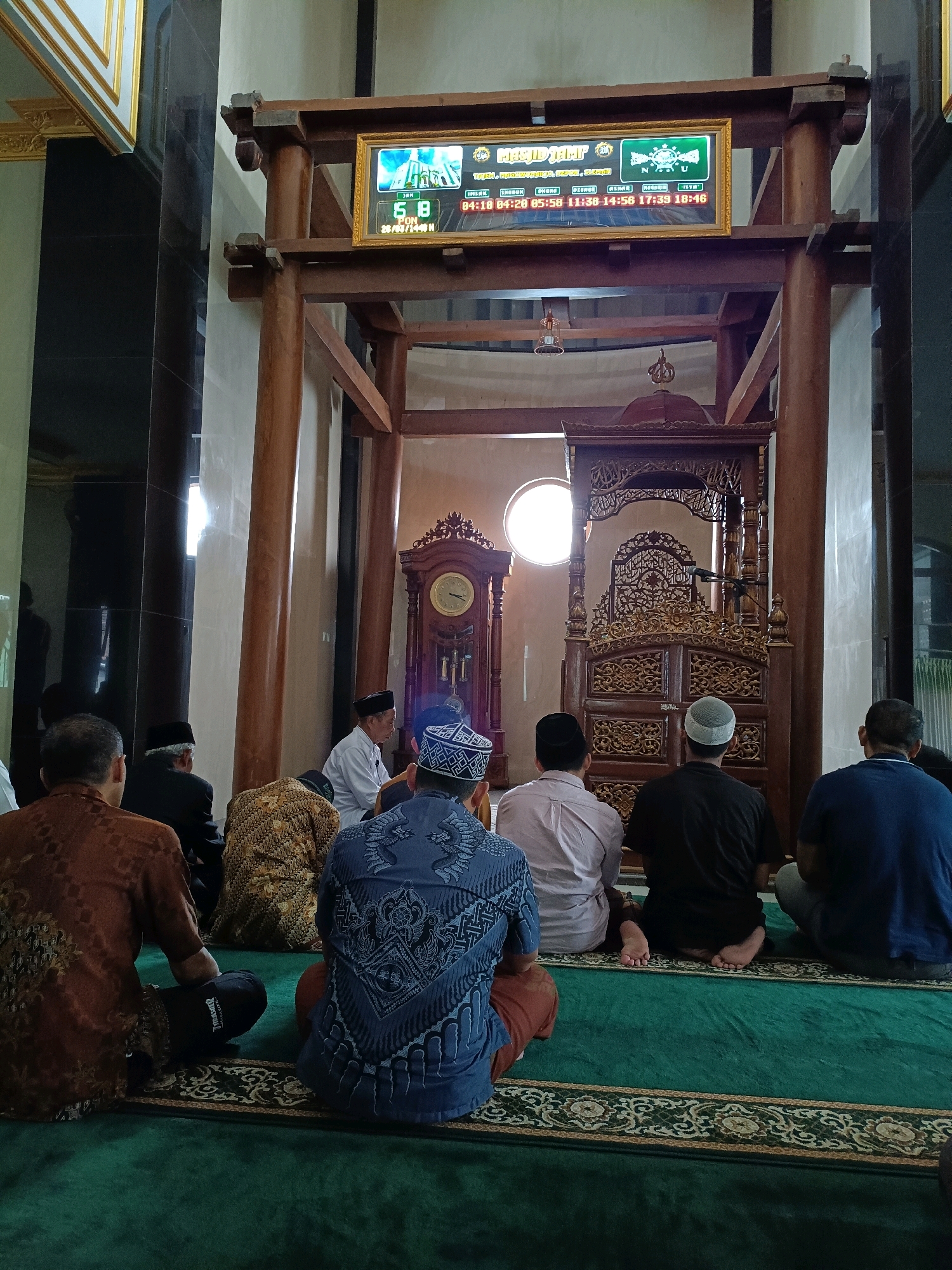 In Maguwoharjo, Indonesia, a group of men are seated on a green carpeted floor within a mosque, facing a wooden pulpit adorned with intricate carvings. A large grandfather clock stands to the left of the pulpit, and a digital display board showing prayer times is mounted above it. The men are dressed in traditional Islamic attire, with some wearing peci (caps). The atmosphere appears to be one of prayer or religious observance. The mosque features tall pillars and a high ceiling.