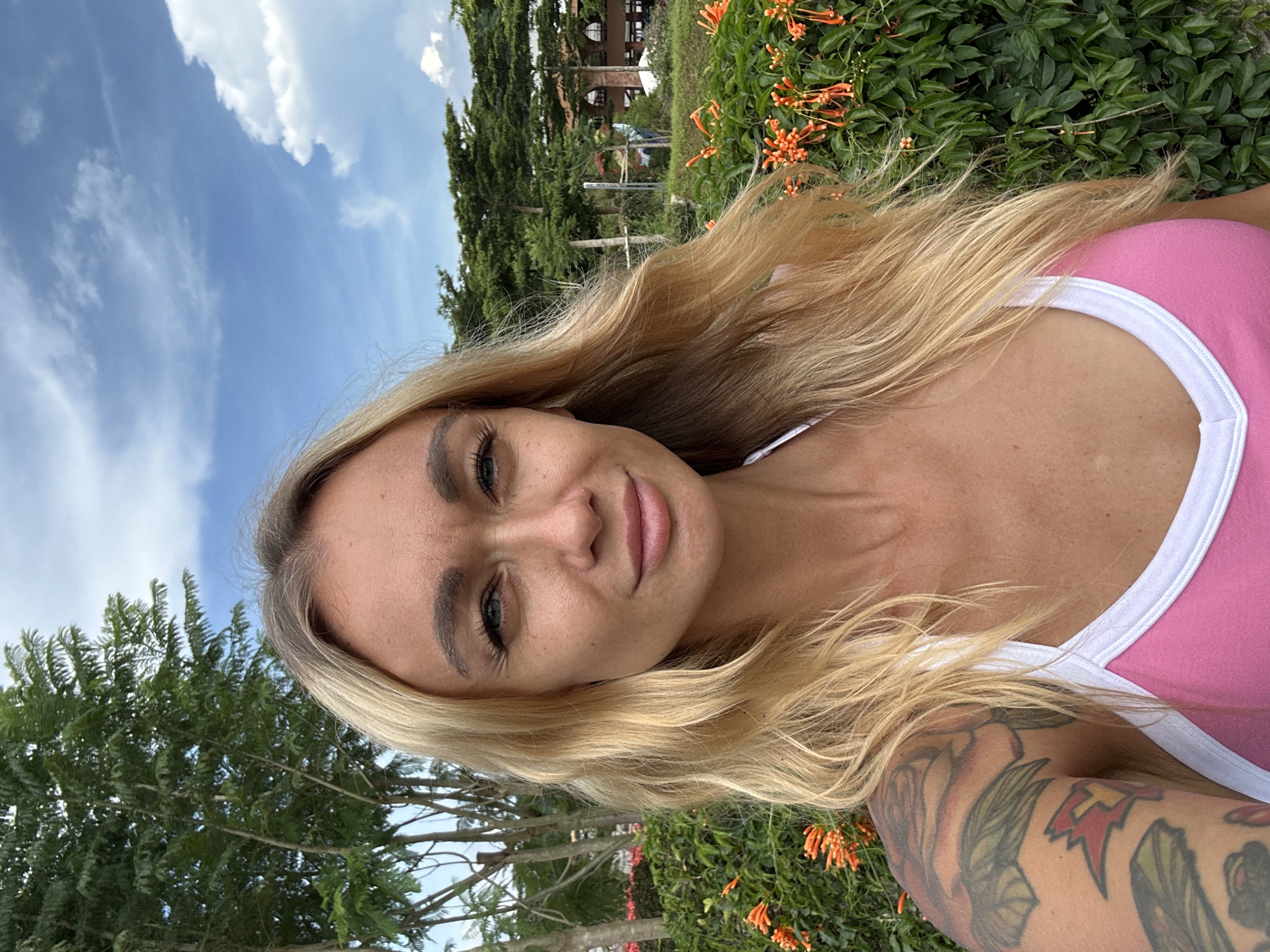 A blonde woman wearing a pink and white athletic top takes a selfie outdoors. She has light blue eyes, dark eyelashes, and full lips. Her blonde hair is long and wavy, framing her face. Her left arm, visible in the foreground, is covered in colorful tattoos.
The background shows lush green foliage, including flowering orange vines, and tall trees. In the distance, a building with a reddish-brown exterior and a tiled roof is partially visible. The sky is a vibrant blue with scattered white clouds. The scene appears to be set during the daytime, likely a warm and sunny day.
The woman's expression is neutral, with a slight tilt of her head as she looks towards the camera. The image has a casual, relaxed feel, suggesting the woman is enjoying her time outdoors. The location context places the scene in Cam Ly, Đà Lạt, Vietnam. No text is visible in the image.