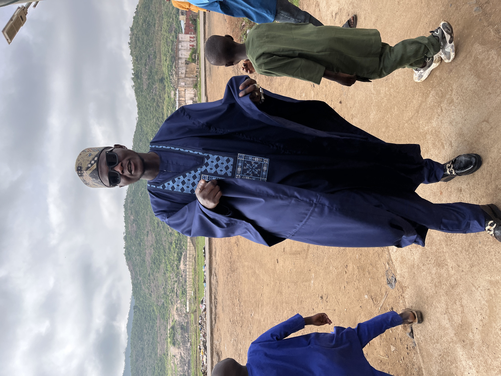 A man in a navy blue boubou and patterned hat stands on a concrete surface in Jos, Nigeria. He wears sunglasses and faces the camera. Other individuals, including children wearing blue and green tops, are partially visible around him. In the background, a mountain is visible under a cloudy sky, along with evidence of urban development and trash.