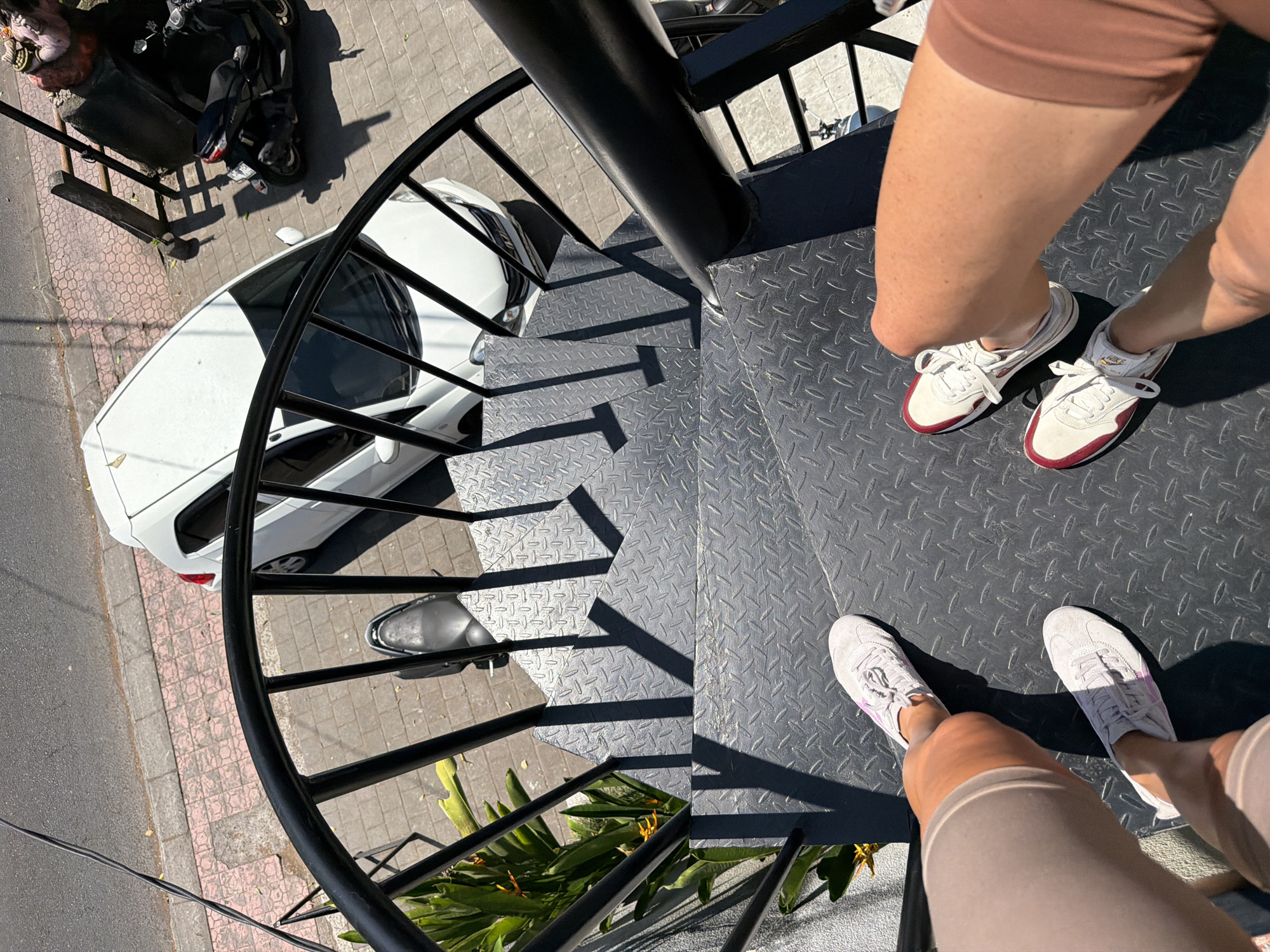 Two individuals are standing on a metal spiral staircase, their legs and feet visible. The staircase has a diamond plate pattern on the treads and a black metal railing. In the background, a white car is parked on a street with a patterned sidewalk. A black scooter is parked next to a statue on the right side of the image. The scene appears to be outdoors, under bright sunlight, casting shadows on the staircase. The location is identified as Ubud District, Indonesia.