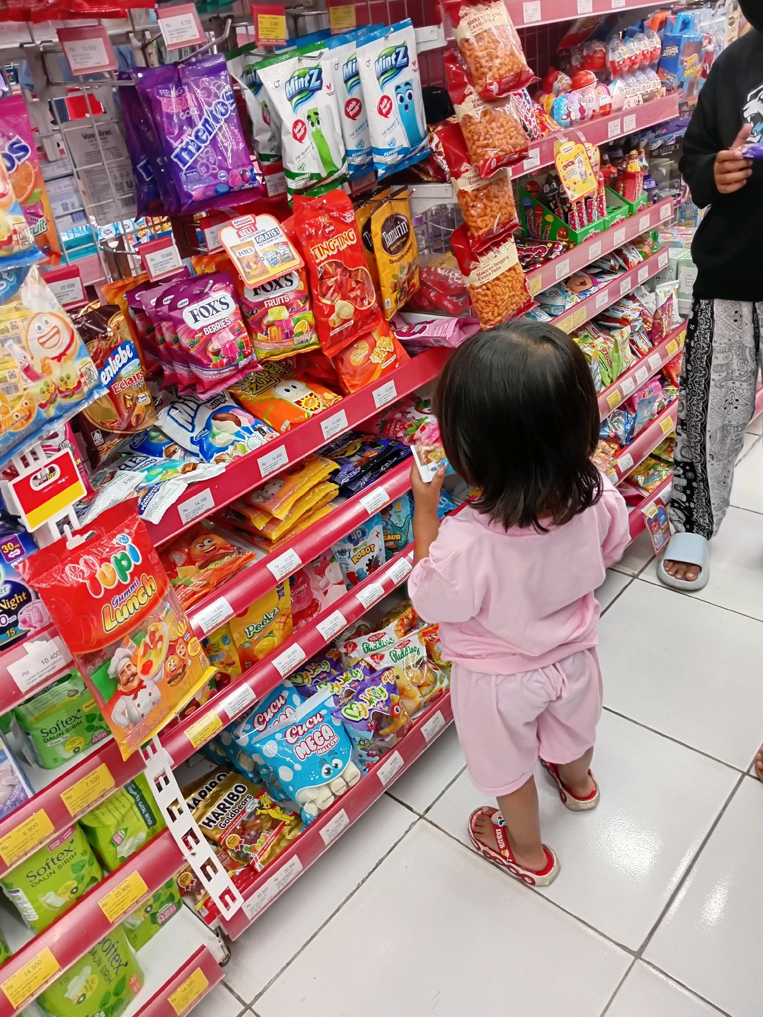 A young child, viewed from the back, wearing a pink t-shirt and shorts, stands in a brightly lit aisle of a supermarket, possibly in Seren, Indonesia. The child is holding a small, white package and is reaching towards a shelf filled with colorful snacks and candies. The shelves are stocked with various brands of packaged goods, including what appear to be gummy candies, biscuits, and chips. The floor is tiled in a white, grid pattern. A person's legs, wearing patterned pants and a sandal, are visible to the right of the child, suggesting another shopper is present. The overall impression is a typical scene of a child browsing snacks in a retail environment.