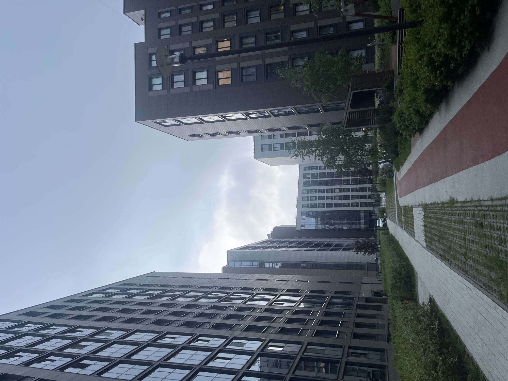 The image shows a view upwards in a courtyard surrounded by high-rise residential buildings, characteristic of Saint Petersburg, Russia. The buildings have a modern design with repetitive windows. A pedestrian walkway runs along the right side of the frame, featuring a combination of concrete pavers, grass, and a red painted section. There is lush green foliage lining the walkway. The sky above is overcast with some areas of visible clouds. No people are visible in the image.