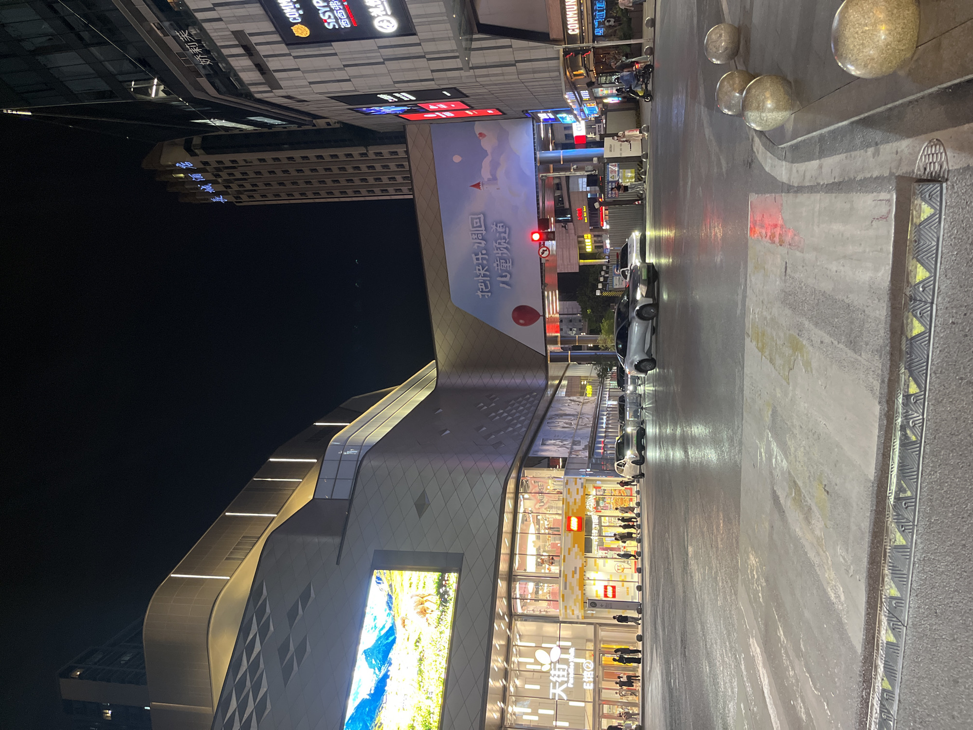 This is a nighttime street-level view of a commercial district in Yuzhong District, China. A multi-story building with a prominent LED screen displaying a mountain landscape is visible on the left. Adjacent to it is another building with large glass storefronts, including a Lego store. To the right, a taller building with a sign reading "长城酒店" (Great Wall Hotel) is illuminated against the dark sky. Further to the right, another building features various illuminated signs, including one for "COMM" and "SiSYP". The street in the foreground is wet, reflecting the lights from the buildings and traffic signals. Several cars are on the road, and a few pedestrians are walking on the sidewalk. The overall scene depicts a bustling urban environment at night, with modern architecture and commercial activity.