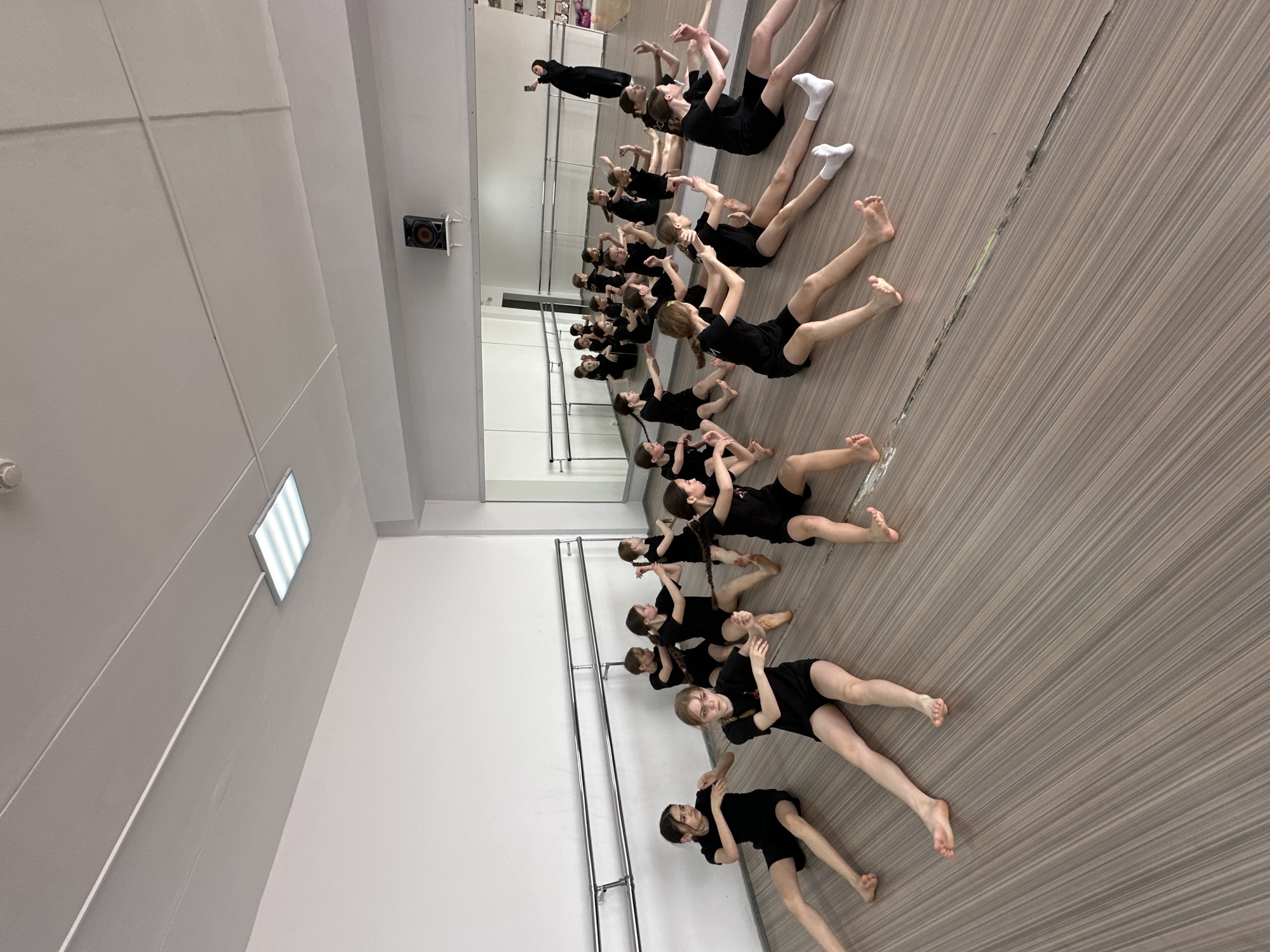 The image shows a dance studio in Votkinsk, Russia. Approximately 25 girls in black leotards are lying on a wooden floor. Some are barefoot, while others wear white socks. They are arranged in rows facing a mirrored wall. The reflection shows the same group and the studio's interior. A barre runs along the wall opposite the camera. The room has white walls with a rectangular light fixture and a speaker mounted high on the wall.