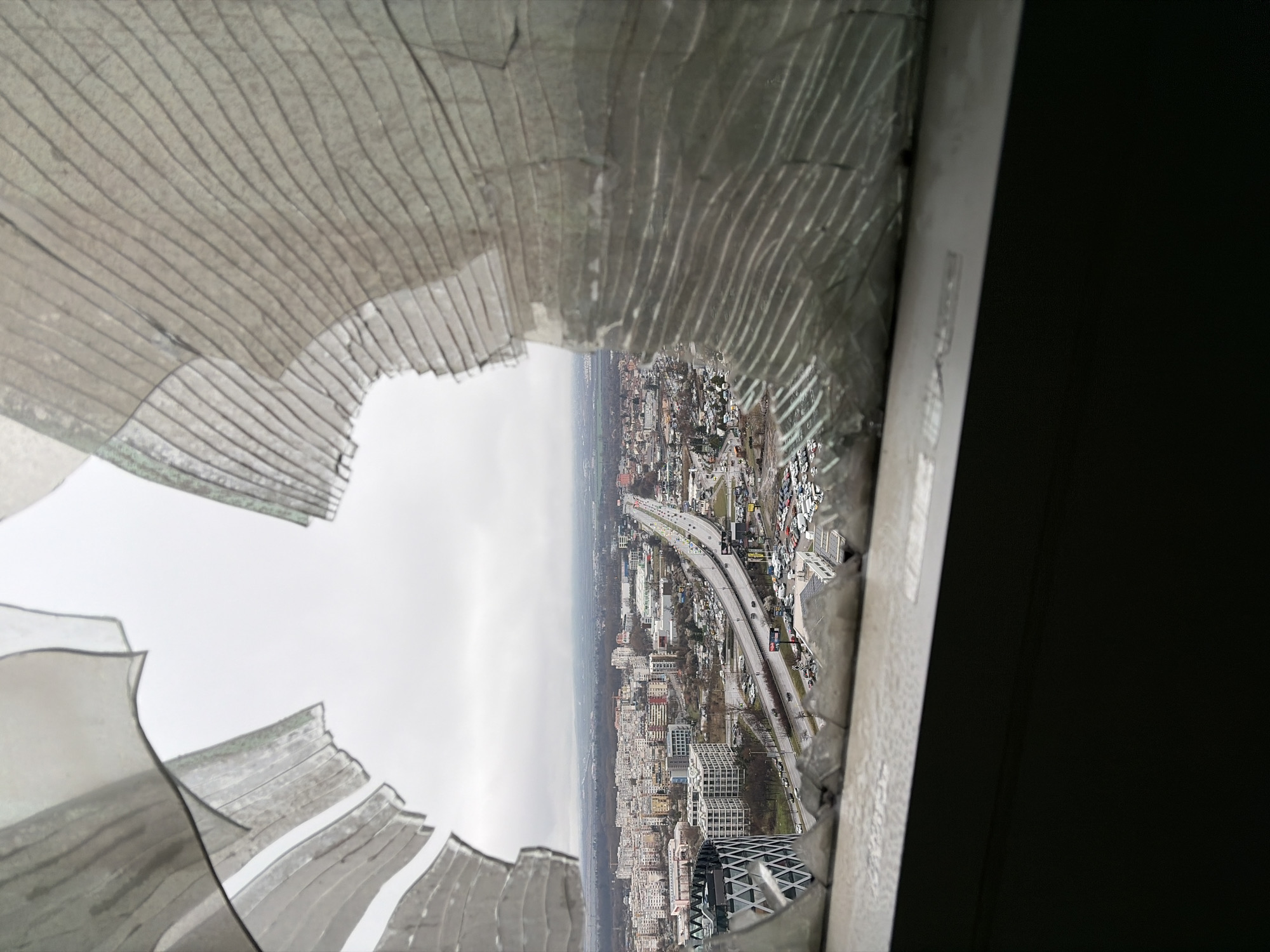 The image presents an elevated view of an urban landscape in Sofia, Bulgaria, observed through a severely fragmented window. The foreground features a window pane constructed of tempered or laminated glass, which has shattered into numerous small, blunt, translucent shards. These shards remain partially attached, forming an intricate network of cracks and creating a large, irregular opening in the upper-center and right portions of the frame. The lower section of the window frame, likely metallic, is visible, with dark, indistinct shapes below it suggesting an interior space.
Beyond the broken glass, the cityscape of Sofia unfolds under an overcast, grey sky, indicating diffuse light conditions. Prominently in the mid-ground, Tsarigradsko Shose Boulevard is visible, a multi-lane highway with an elevated section or overpass where vehicles are observed in transit. To the left of the highway, the distinctive, modern Capital Fort building, characterized by its geometric white and black facade, stands partially visible. Surrounding structures include numerous multi-story residential and commercial buildings, varying in architectural style. Several parking lots are discernible, one notably filled with a high density of white vehicles, potentially indicating a dealership or a large corporate parking facility. The ground, especially on roads and parking areas, appears wet, consistent with rain or damp conditions. No discernible individuals or specific interactions are visible due to the distance. The environment suggests an urban setting on a non-summer day.