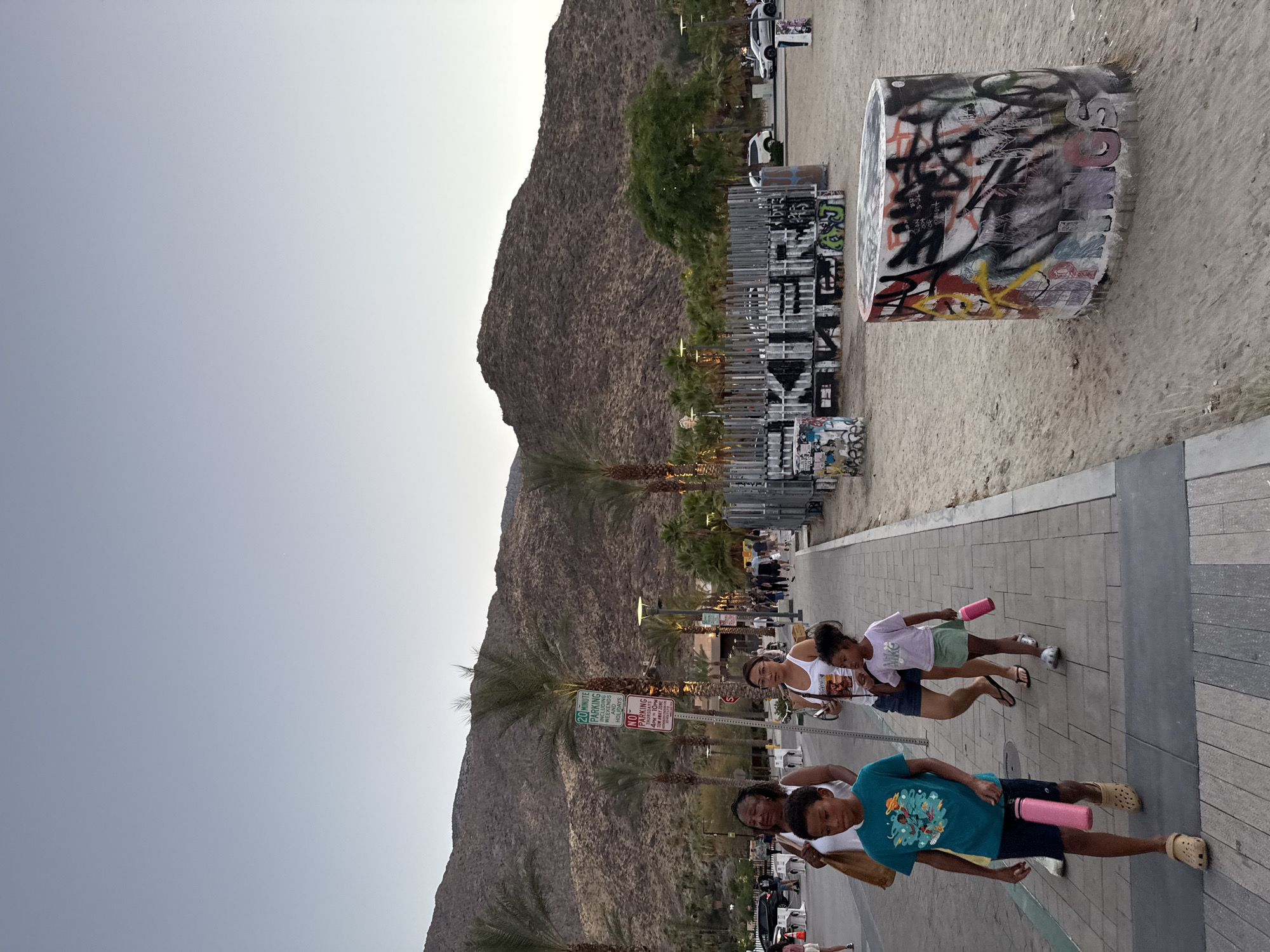 In Palm Springs, California, USA, a family walks along a paved walkway, which borders a sandy area. In the background, a mountain range dominates the horizon, with sparse vegetation and palm trees dotting the landscape. A metal structure, covered in graffiti, stands to the right of the walkway. In the foreground, a cylindrical concrete planter is also adorned with vibrant graffiti. The sky above is a clear, pale blue, suggesting either early morning or late evening.
